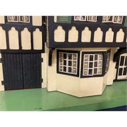 Large Tri-ang No.93 doll`s house in the 'Stockbroker's Range' c1935; two-storey double-fronted Tudor design, half timbered gables, two-storey bay windows, cream and mock shrubbery to front, opening metal framed windows with green shutters, red simulated tiled roof with chimney, integral garage with opening doors, front elevation has four hinged doors opening to reveal two bedrooms with fire places, living room, bathroom and kitchen with built in dresser and sink, entrance hall and staircase, side entrance with porch and seat and sun dial over on chimney stack; some wiring for electrical lighting; metal Tri-ang Toys disc verso; comprehensively furnished H66cm L117cm D43cm