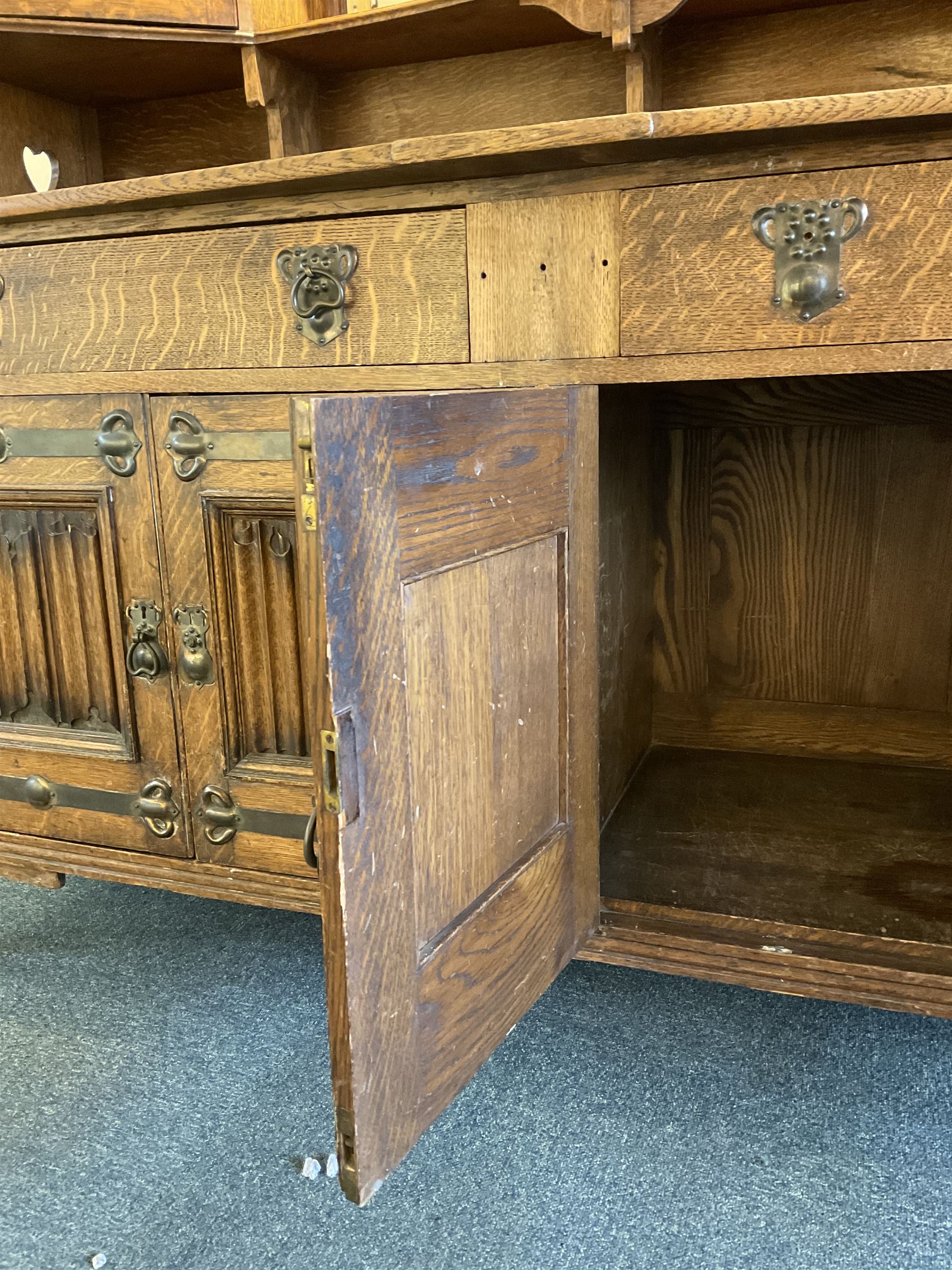 Shapland and Petter - Arts & Crafts period oak dresser, raised gallery top with spade balustrade, central bevelled mirror flanked by two cabinet, astragal glazed doors with yellow tinted glass and bullseye panes, the sides with pierced heart shaped decoration, fitted with two drawers and two double cupboards, enclosed by linenfold panelled doors,  copper work strap hinges and fixtures, on shaped spade feet, the locks stamped 'S&P'