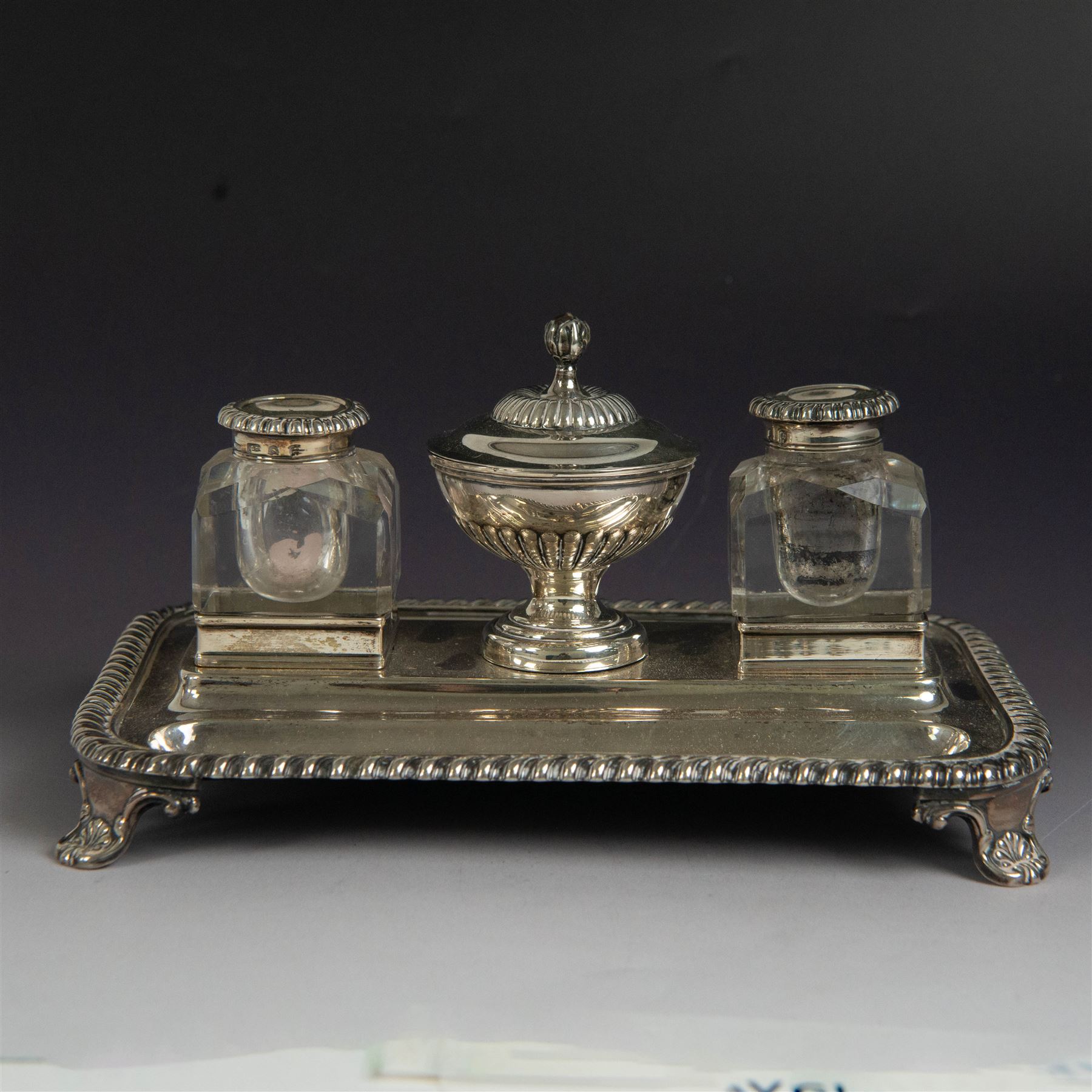 George IV silver desk stand, of rectangular form with rounded corners and oblique gadrooned rim, upon four embossed pad feet, with central pedestal dish of part fluted form with domed lid engraved with crest and with bud finial, with two recesses either side housing faceted glass inkwells, each with silver collars, hallmarked John & Thomas Settle, Sheffield 1825 & 1827, H11cm