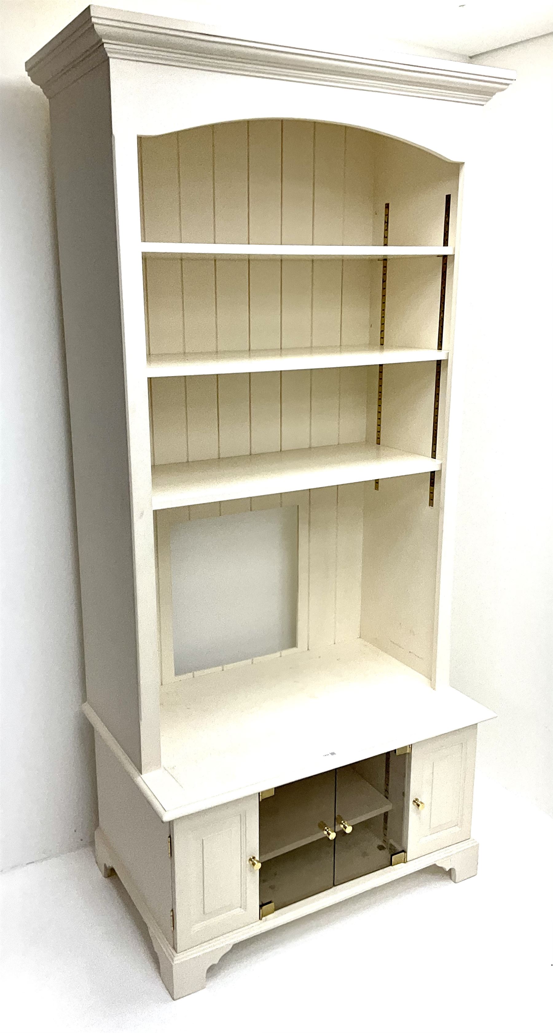 Cream painted bookcase on media stand, projecting cornice, three adjustable shelves above two glazed doors flanked by two cupboards, bracket supports 