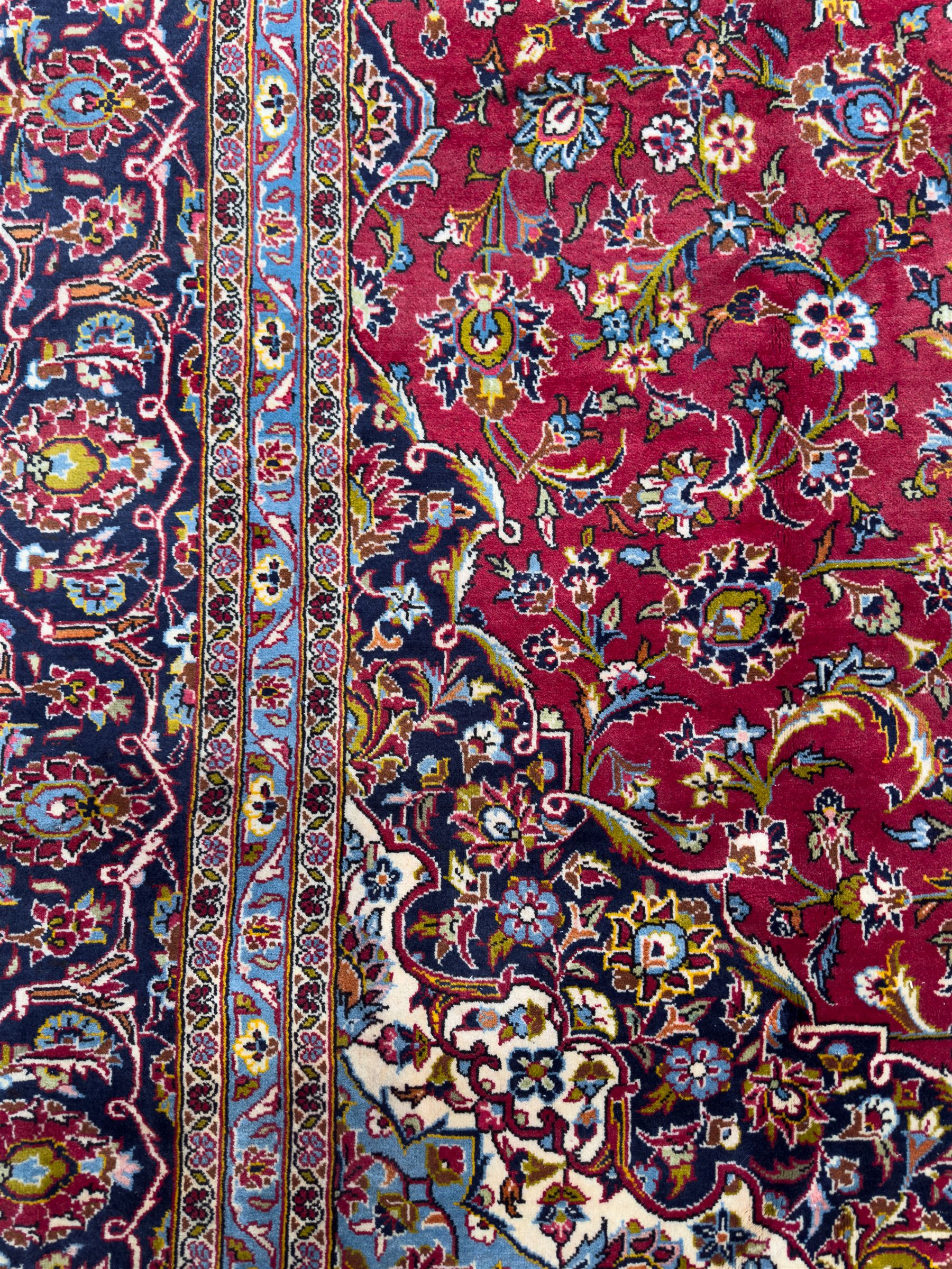 Central Persian Kashan hand-knotted carpet, the madder red field with central lobed medallion in indigo and ivory, enclosing scrolling arabesques and flowerheads, framed by palmette spandrels and an indigo border of rosettes and lotus forms between multiple floral guard stripes, from the region of Kashan
