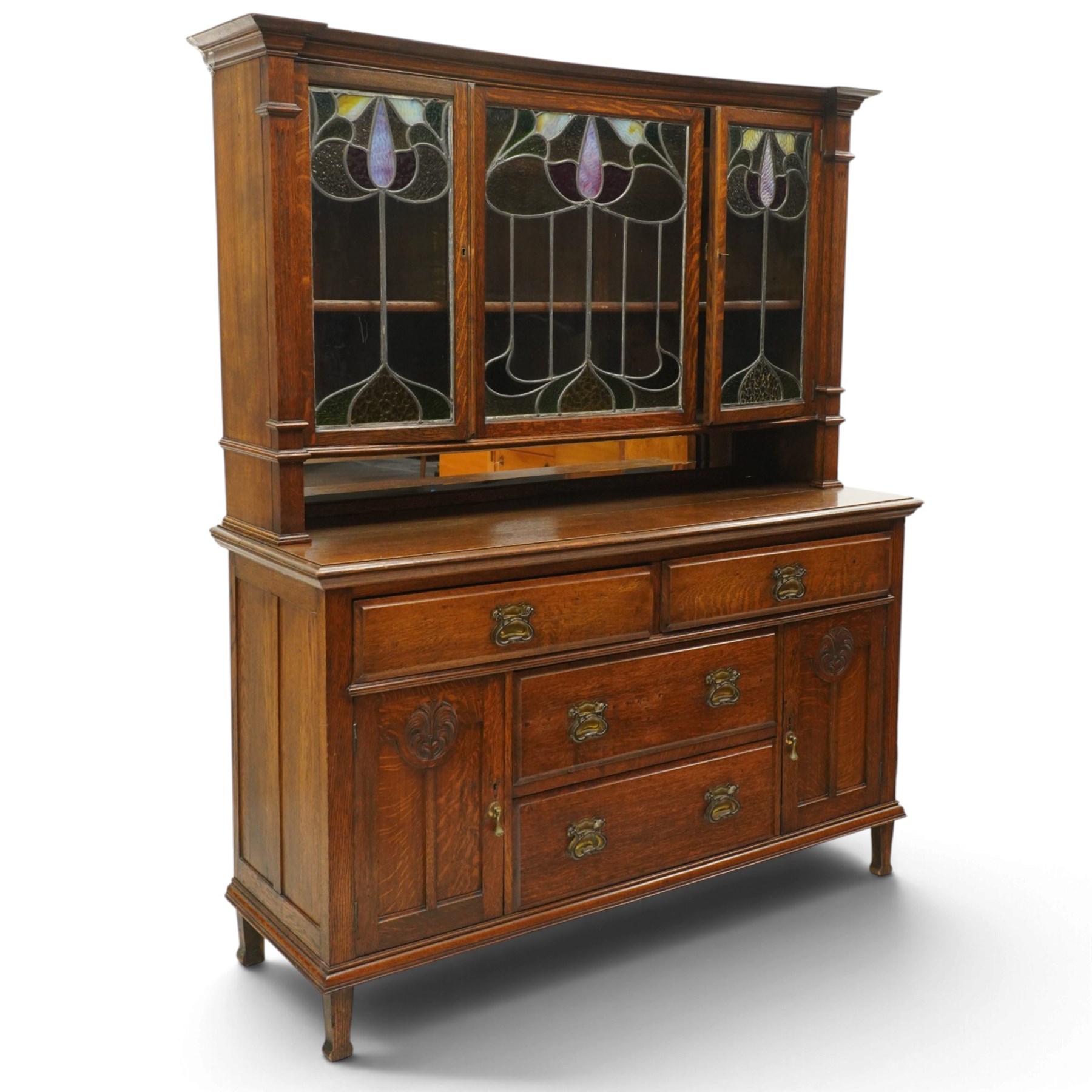 Arts and Crafts oak dresser, projecting moulded cornice above three glazed panels with coloured leaded glass floral panels, flanked by two glazed doors enclosing two adjustable shelves, open mirrored recess below with mirrored back, the base fitted with two short drawers over two central drawers flanked by carved panelled cupboard doors, ornate brass handles, raised on tapering square legs