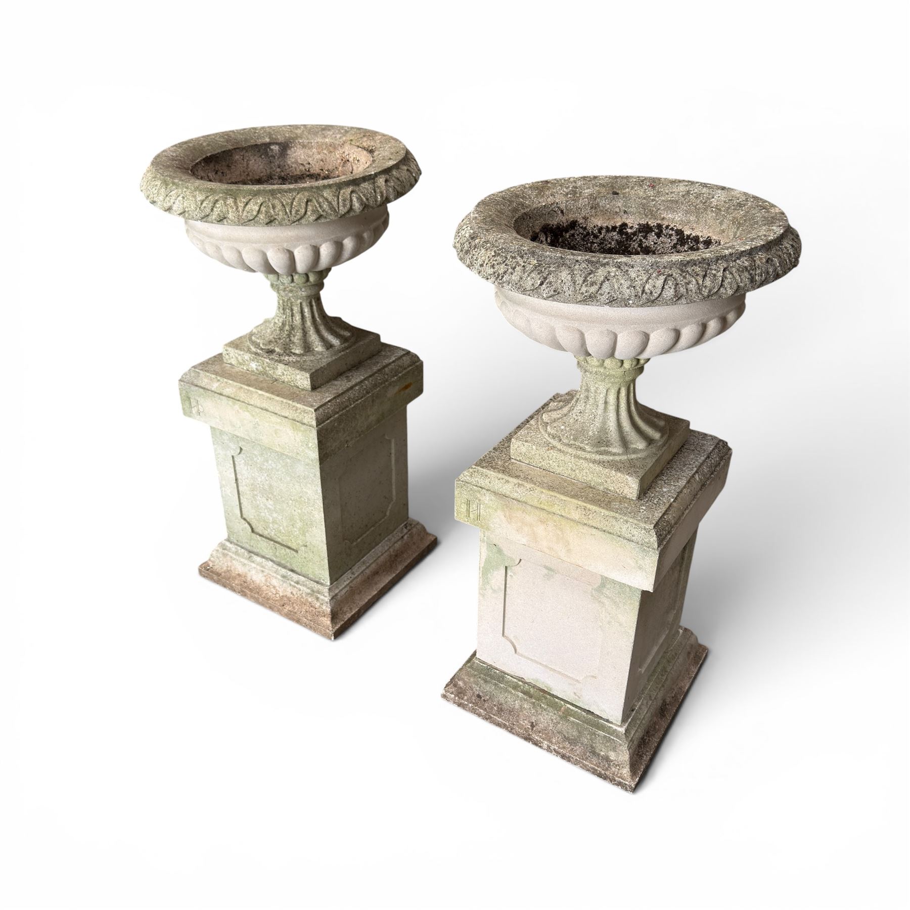 Pair of weathered cast stone Neoclassical design garden Kylix shape urns, foliate moulded rim over a gadrooned underbelly, the scalloped socle over a rectangular pedestal with faux-panelled sides, stamped with stylised ‘H’ monogram