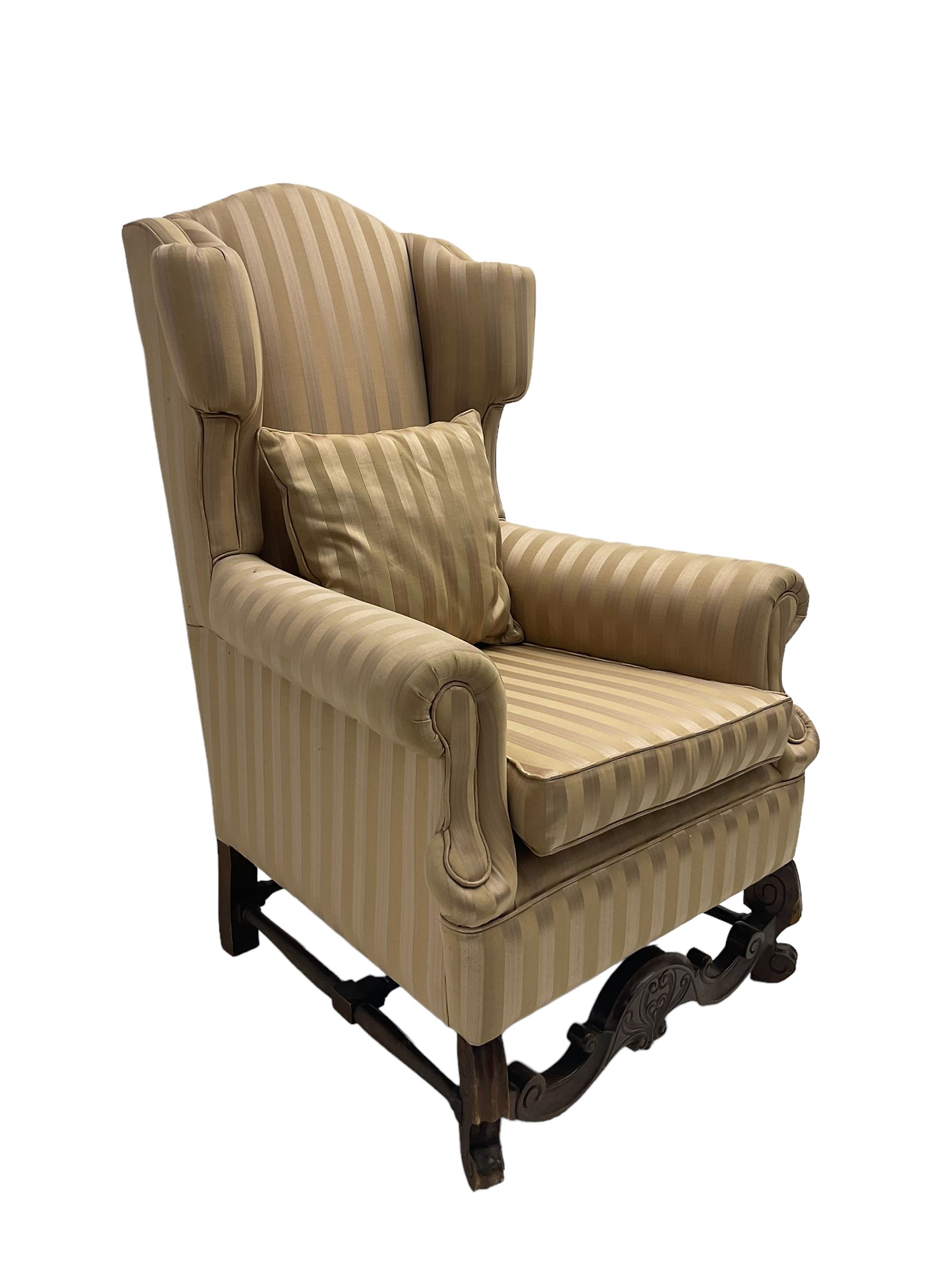William and Mary style wingback armchair, upholstered in gold striped fabric, on walnut scroll carved supports joined by scroll and foliate carved middle rail, turned H-shaped stretchers, on castors