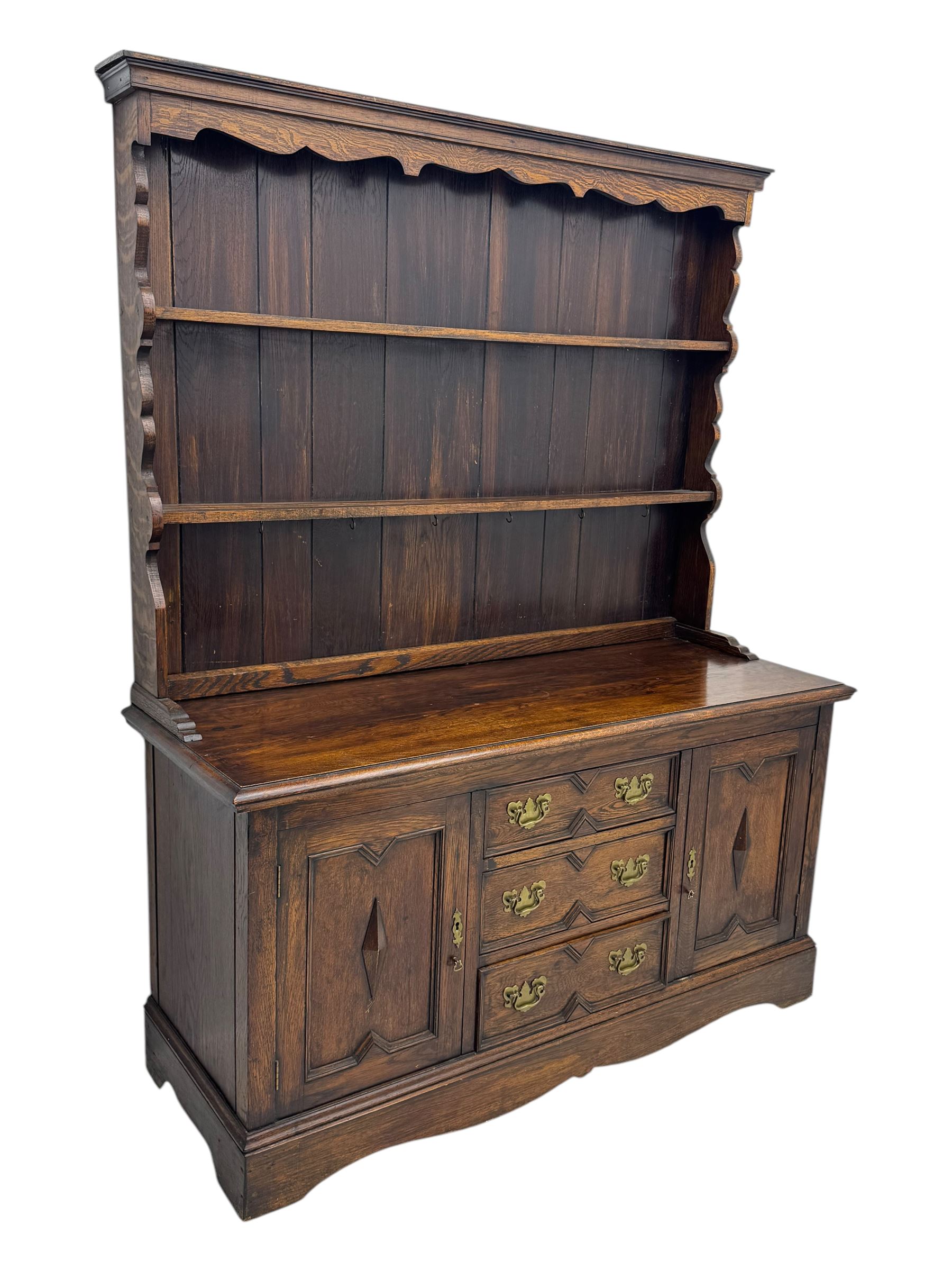 Late 19th century oak dresser, the upper section with two shelves beneath a shaped cornice, the lower section with a central bank of three drawers flanked by cupboard doors, all decorated with geometric diamond motifs, the drawers fitted with brass handles, raised on a shaped plinth base
