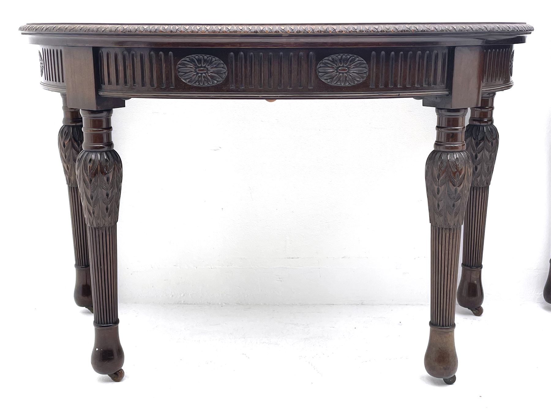 Pair early 20th century Hepplewhite style mahogany D-end console tables, the curved top with foliate carved edge, fluted frieze rails carved with flower heads, turned and reeded acanthus supports with recessed castors