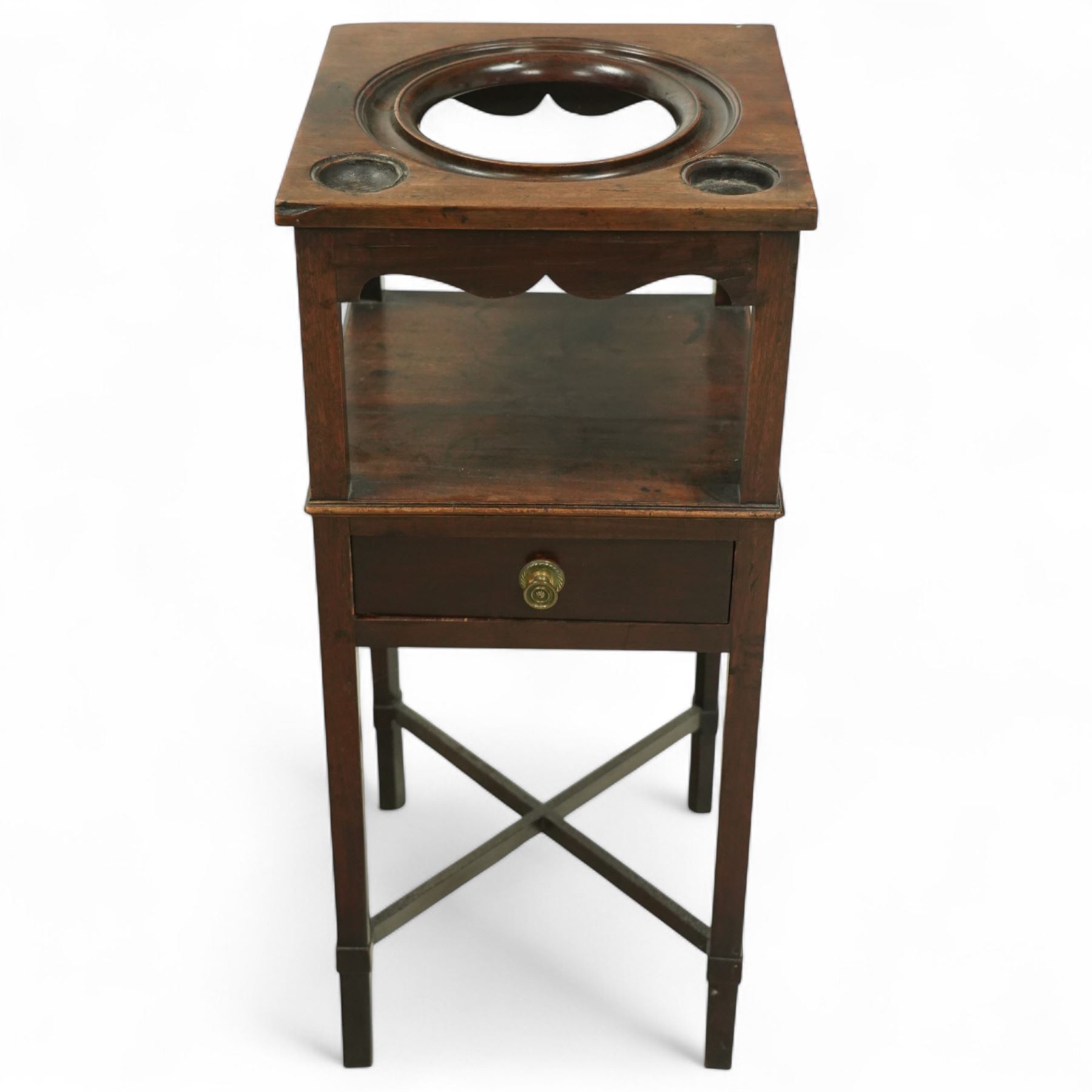 George III mahogany washstand, square top with circular wash bowl aperture and twin bottle recesses above a shaped frieze and single drawer with brass knob handle, raised on square tapered legs united by cross stretchers 

