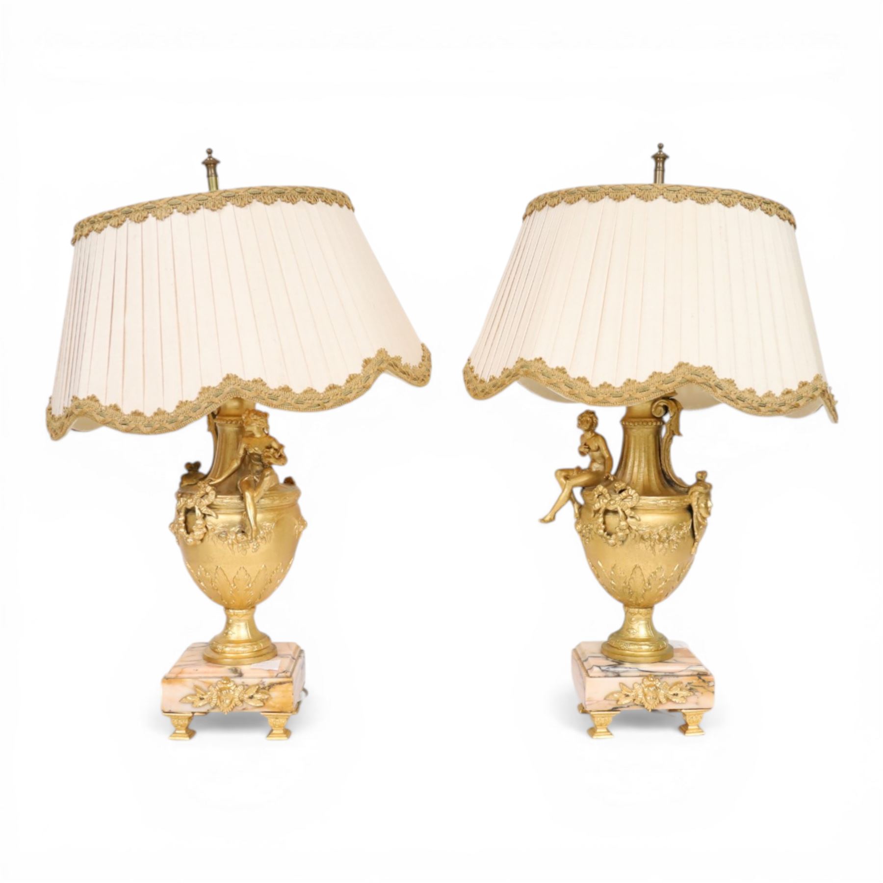 Pair of Rococo style gilt metal table lamps, the urn shaped bodies each with applied figure seated upon shoulder, and female mask to opposite side issuing scroll leading to top of neck, the body cast and applied with stiff leaf detail, and foliate swags and wreaths, upon square pink marble plinths with applied gilt metal mask mounts, and four gilt metal bracket feet, each supported a pleated cream silk closed top lampshade, H63cm