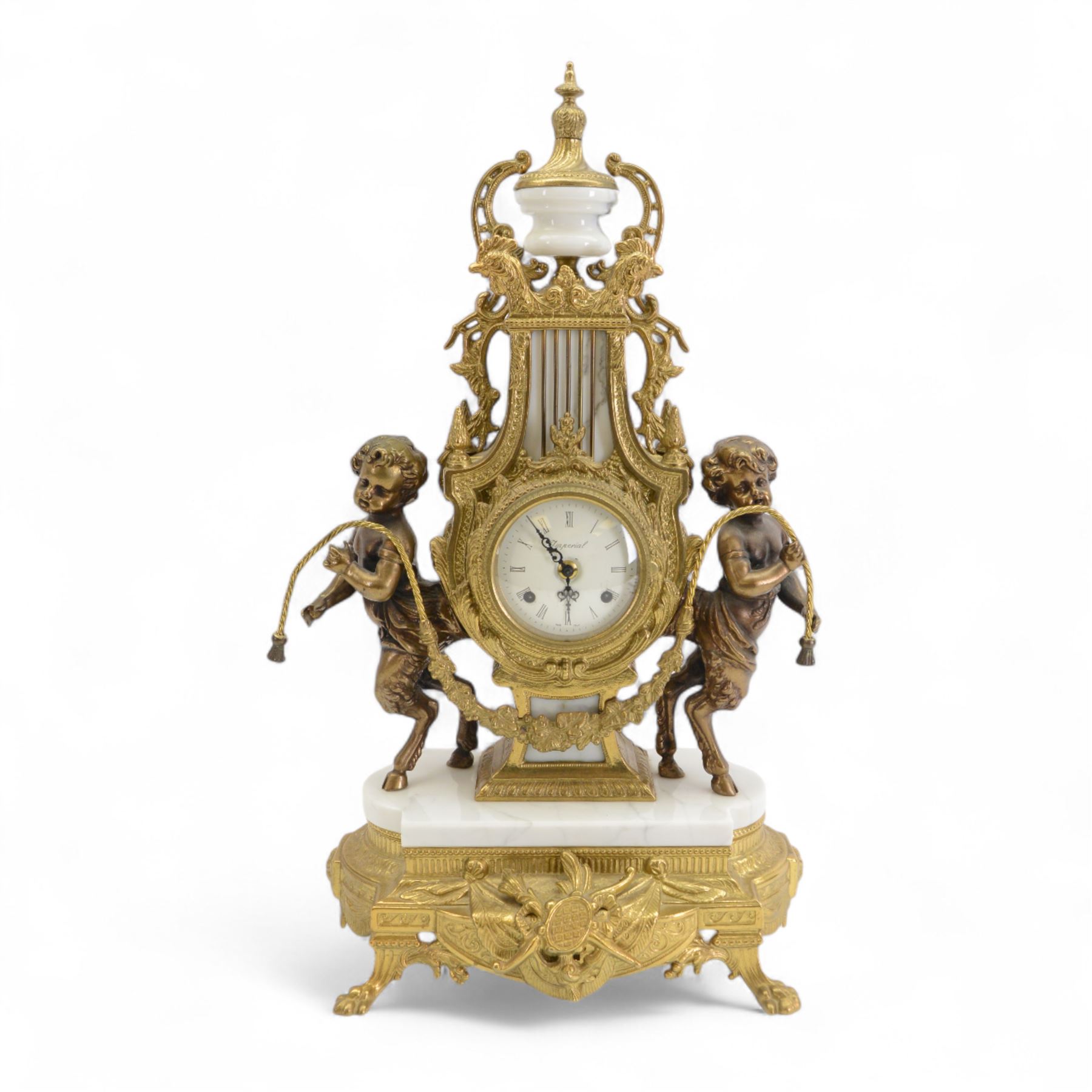 Italian- 20th century 8-day gilt figural mantle clock in the style of a 19th century Parisian lyre clock, drum movement with a porcelain dial flanked by playful cloven-hoofed cherubs on a white marble plinth, raised on a a decorative base with splayed paw feet, twin train spring driven Hermle movement with a floating balance escapement, sounding the hours and half hours on two bells. With key.    