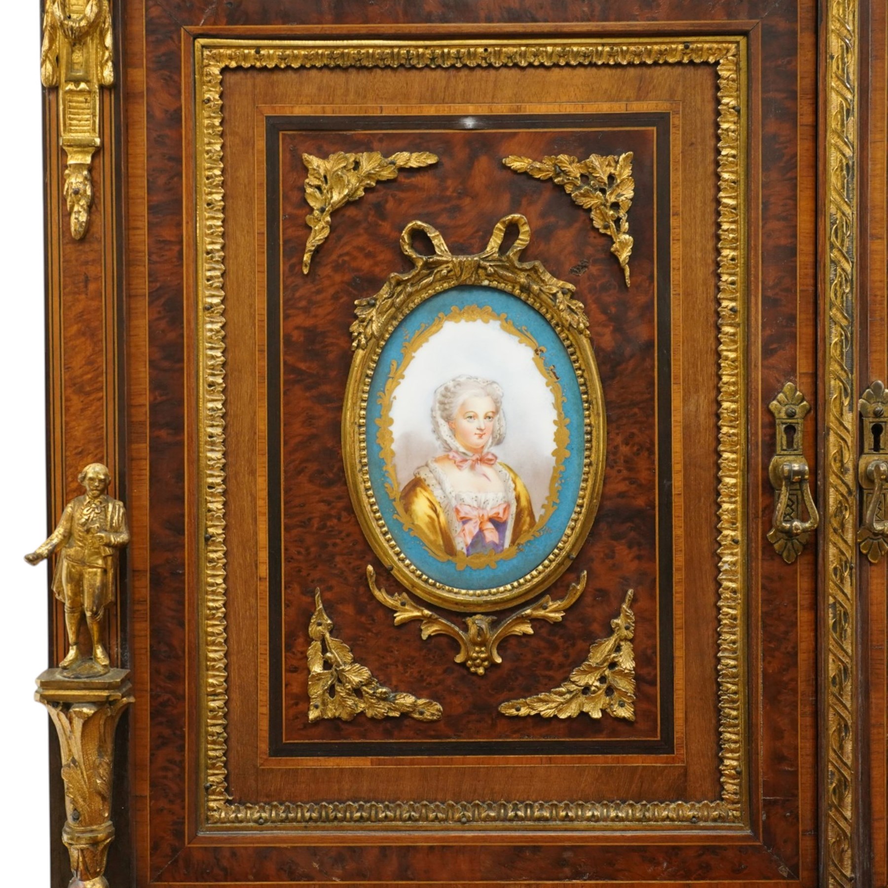 Fine late 19th century Aesthetic Movement ebony and amboyna cabinet on stand with parcel-gilt detail, the moulded cornice with pierced gilt-brass three-quarter gallery above a pair of panelled doors centred by oval porcelain portrait plaques within gilt-metal ribbon-tied frames, the doors further enriched with gilt-metal foliate mounts and flanked by classical figural terms, enclosing a shelved interior, the projecting lower section with amboyna top above a frieze drawer set with a series of floral porcelain roundels within pierced gilt-metal mounts and a central oval floral plaque, raised on turned and fluted supports with gilt-heightened detail, the mirrored back behind two amboyna shelves with pierced gilt-brass galleries, on turned feet