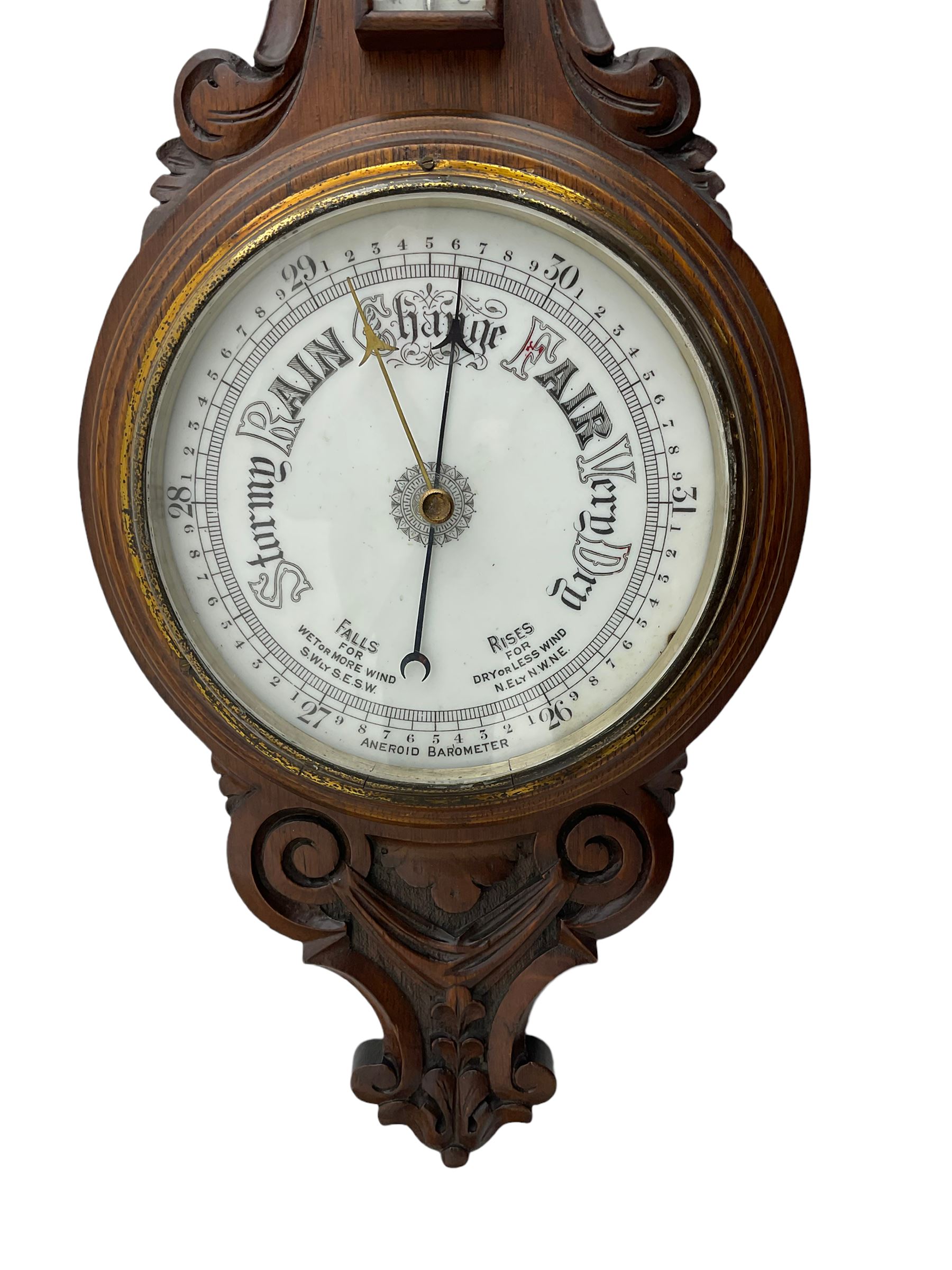 An English 1930’s solid oak carved hall barometer in a scroll shaped carved case with relief carving, aneroid movement, 8” white register measuring barometric air pressure from twenty-six to thirty-one point nine inches, weather predictions in black upper and lower case gothic script with a blue steel indicating hand and brass recording hand, brass dial bezel with flat bevelled glass, mercury thermometer enclosed in a glazed rectangular box displaying temperature in degrees Fahrenheit and Celsius on a white register. H87cm


