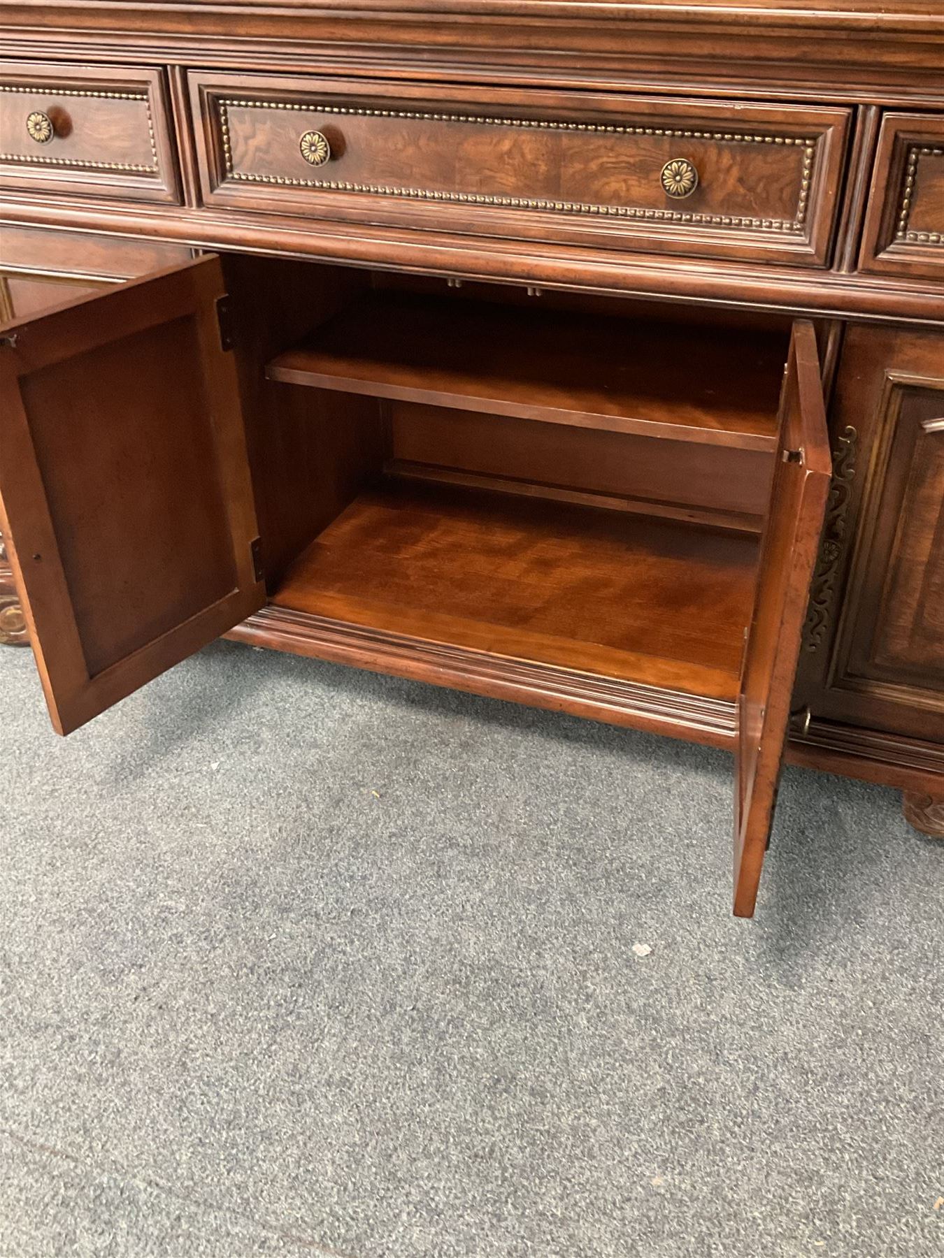 Kevin Charles American walnut display cabinet, illuminated interior, shell carved pediment above four bevel edge doors enclosing six glazed shelves above one central long drawer flanked by two short drawers, above three cupboards, carved bracket supports 