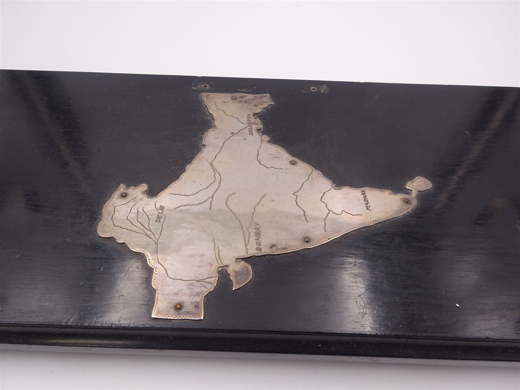 Late 19th/early 20th century Indian silver scroll holder, of cylindrical form with vacant cartouche and embossed with figures, animals and huts, upon a shaped ebonised stand with silver supports and silver panel of India