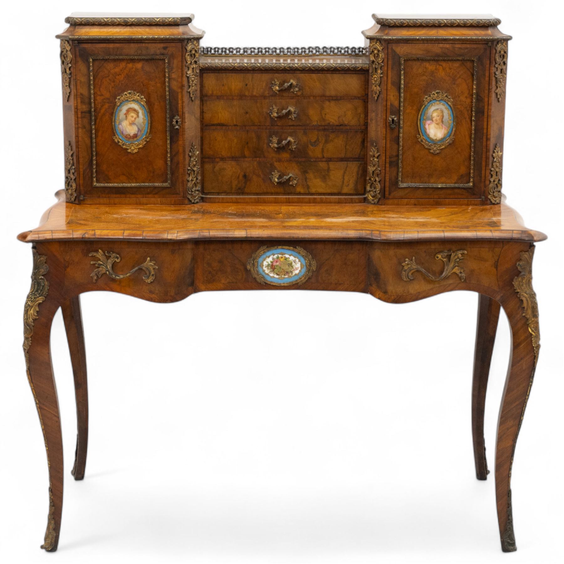 Late 19th-century Louis XV revival gilt bronze mounted and walnut Bonheur-de-Jour, rectangular-shaped serpentine form, with gilt bronze beading, frieze with two blind drawers and gilt bronze handles, centre drawer with large central serves style porcelain plaque, the elevated section with brass gallery above panelled cupboard doors also with serves style porcelain plaques and gilt bronze mounts, interior with single shelf, central section with four drawers each with gilt bronze handles, raised on slender cabriole legs fitted with gilt bronze rococo style masks and sabots