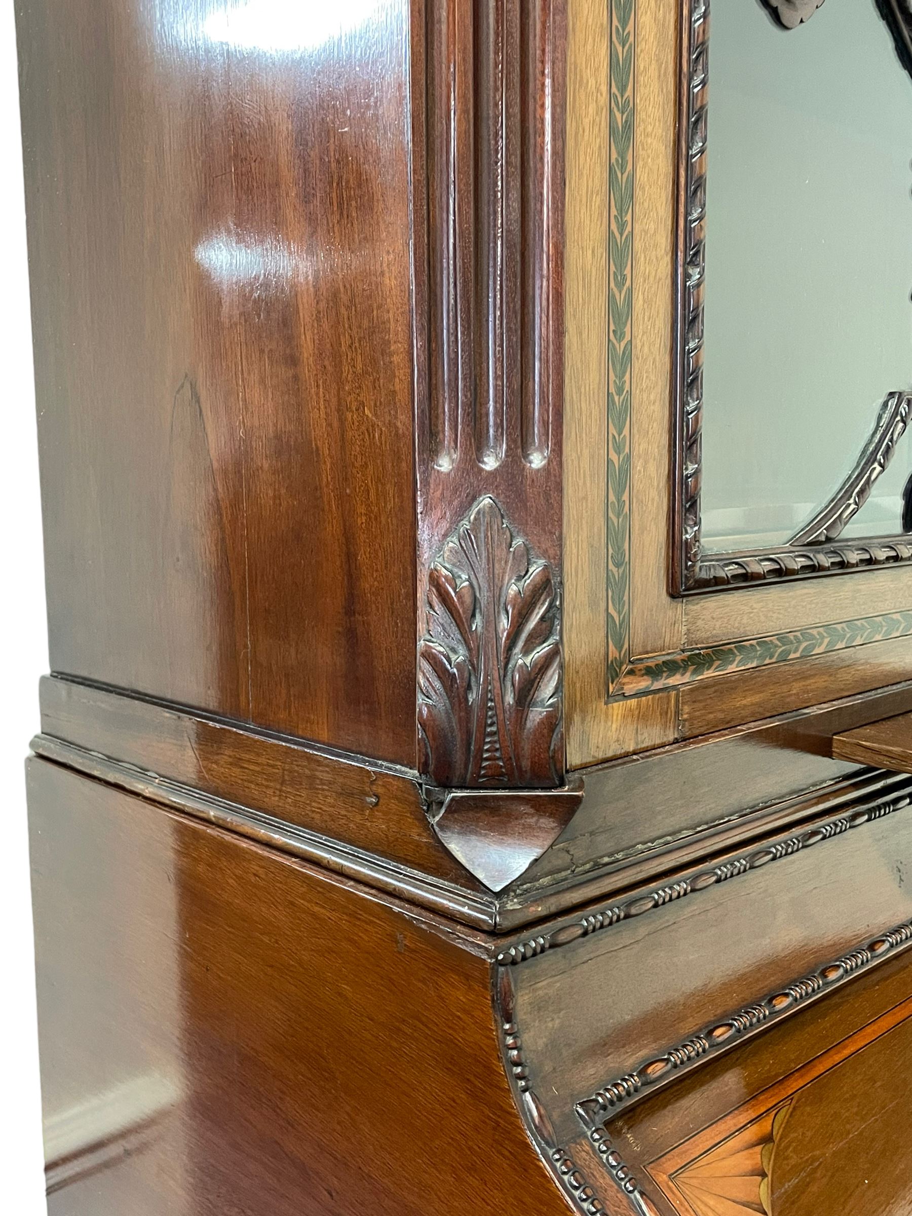 Large and impressive late 19th century Irish mahogany secretaire bookcase, moulded sloped arch dentil pediment decorated with central carved mask surrounded by linen festoons and trailing bellflowers, canted and fluted uprights with carved acanthus leaf upper and lower brackets, enclosed by two astragal glazed doors in the form of trophies with twist ribbon carved mouldings, inlaid with fanned spandrels and trailing leaf painted satinwood band, cylinder roll top inlaid with central fan motif and fanned spandrels, pull-out action revealing stepped fitted interior, central tambour roll cupboard flanked by two Composite pilasters and a combination of drawers and pigeon holes, fitted with three break-front drawers, matched canted and fluted corners with acanthus leaf carvings, on square tapering and fluted spade feet