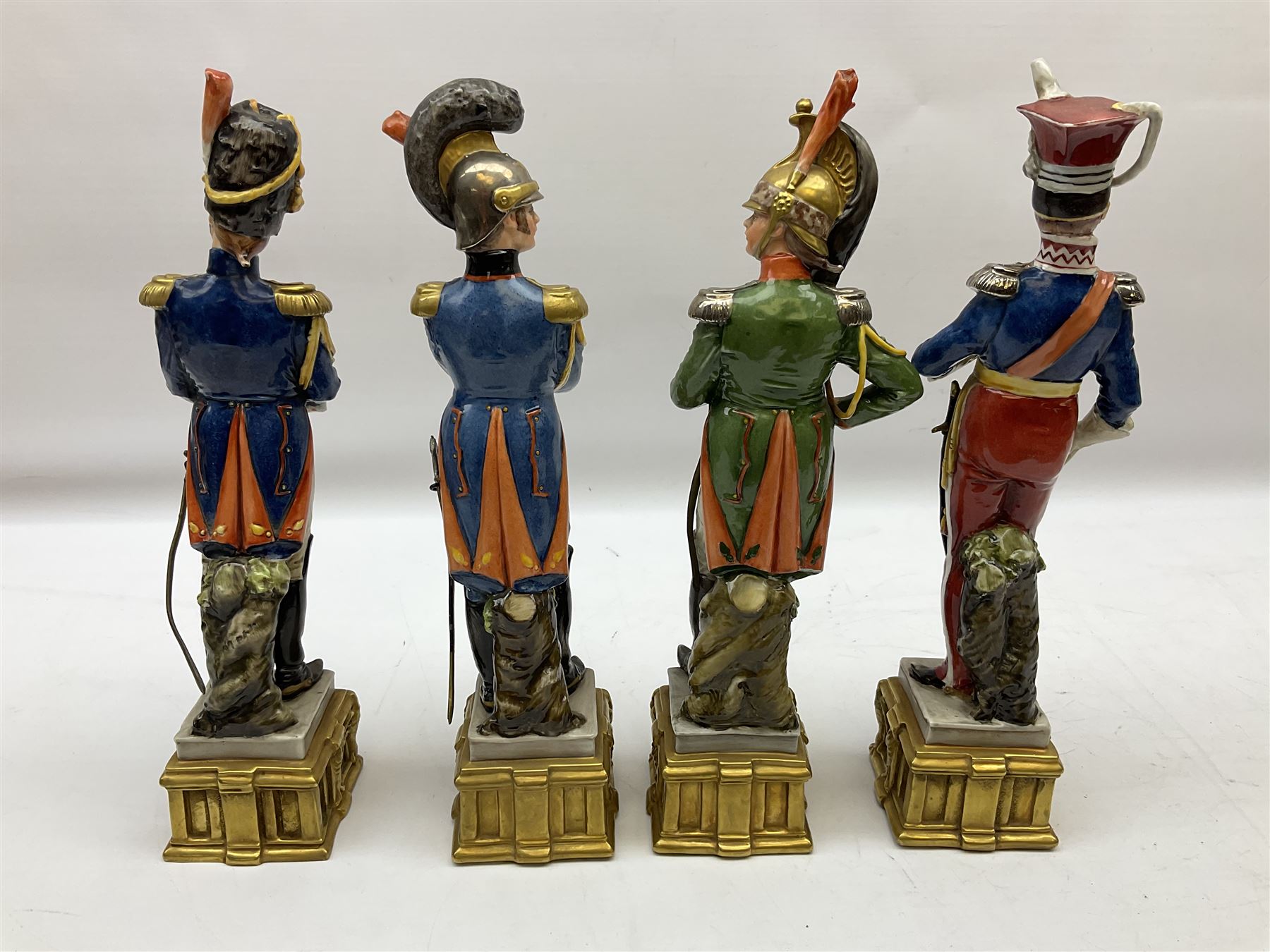 Group of seven Capodimonte figures of soldiers, to include four Bruno-Merli examples including Officer of the 12th Lancers 1820 and Captain of the 18th Hussars 1815, both raised upon ornate gilt plinths bearing British monarch motto in Latin 'Dieu et mon droit', and three further similar, all with crowned N mark beneath, tallest H32cm