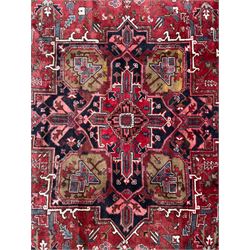 Northwestern Persian Heriz hand-knotted carpet, madder red field with a bold stepped medallion in indigo, ivory, and ivory, surrounded by angular floral motifs and stylized leaves, and the spandrels set within a dark border of rosettes and angular vine motifs, 335cmx252cm