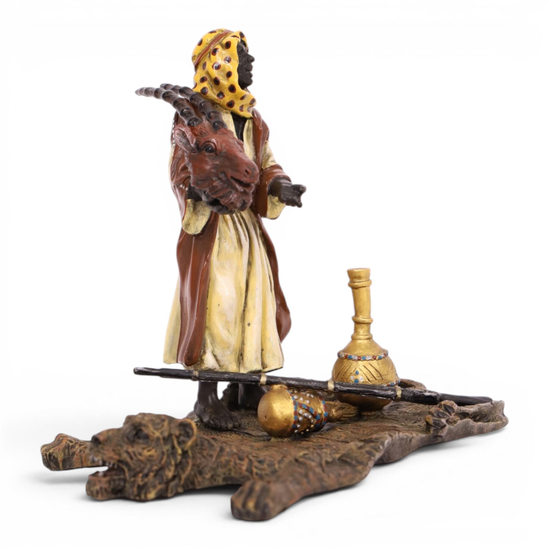 After Bergman - bronze Arab trader holding an antelope head while standing on a tiger skin, with a rifle and jars at his feet, H14cm