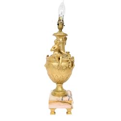 Pair of Rococo style gilt metal table lamps, the urn shaped bodies each with applied figure seated upon shoulder, and female mask to opposite side issuing scroll leading to top of neck, the body cast and applied with stiff leaf detail, and foliate swags and wreaths, upon square pink marble plinths with applied gilt metal mask mounts, and four gilt metal bracket feet, each supported a pleated cream silk closed top lampshade, H63cm