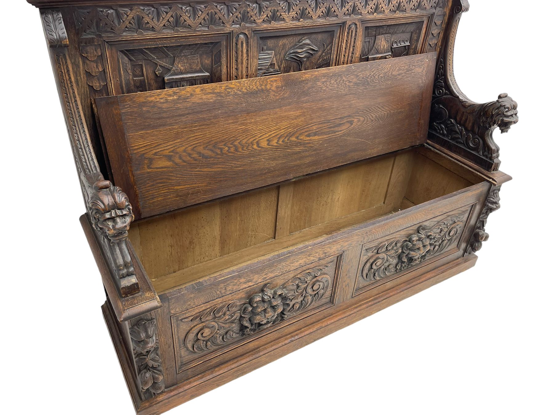 19th century heavily carved oak settle, the moulded upper rail over triangular foliage carved frieze, three Flemish style carved panels depicting tavern scenes, acanthus carved uprights terminating to lion carved arms, hinged box seat, the panelled front carved with lion masks and extending scrolled acanthus leaves, on moulded skirt base