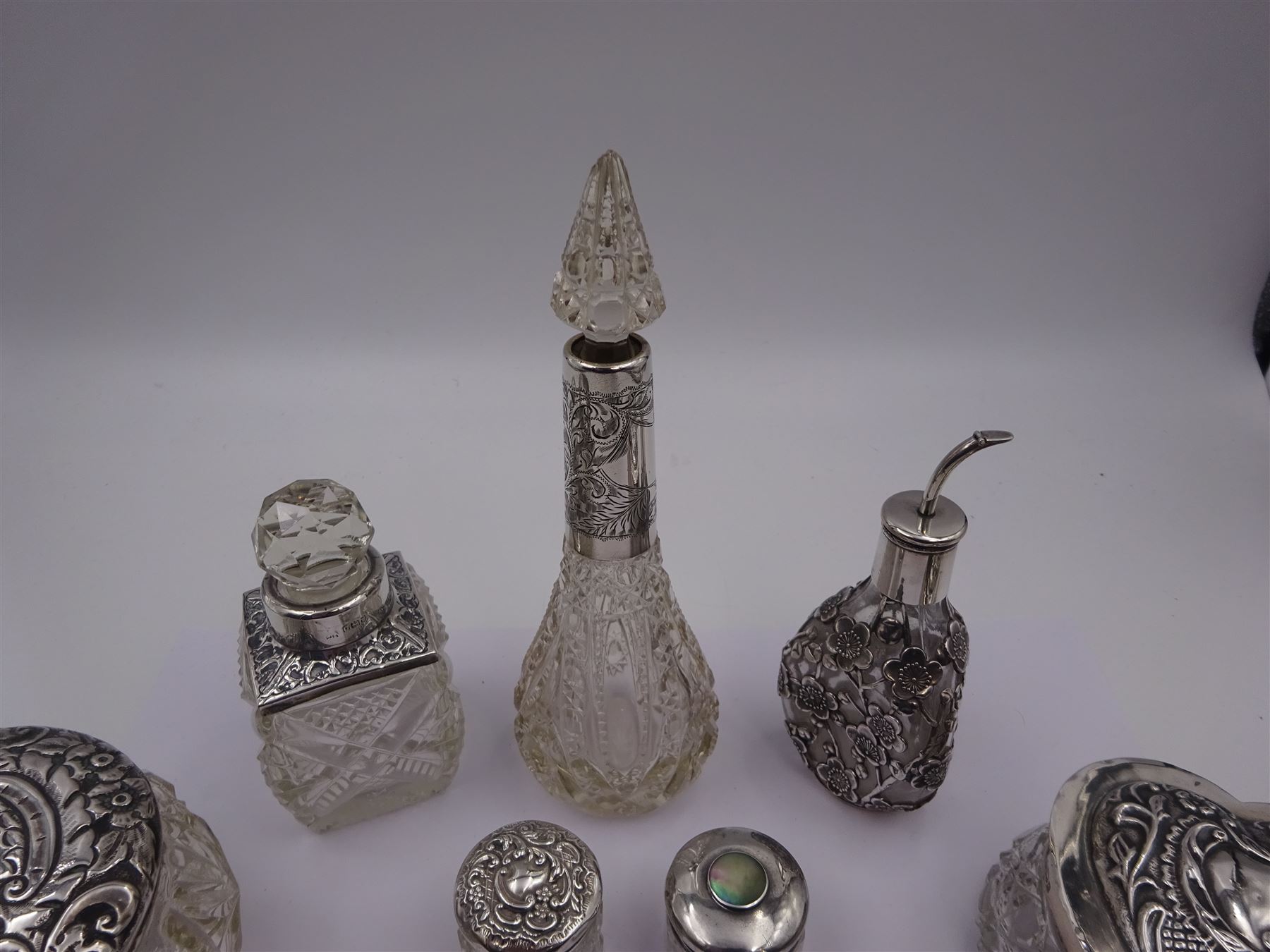 Two silver mounted dressing table jars, one in the form of a heart, both with embossed silver covers and cut glass bodies, hallmarked, together with two smaller examples, one set with mother of pearl, three silver mounted glass scent bottles, including an example with prunus blossom decoration, stamped Sterling, tallest bottle H17cm