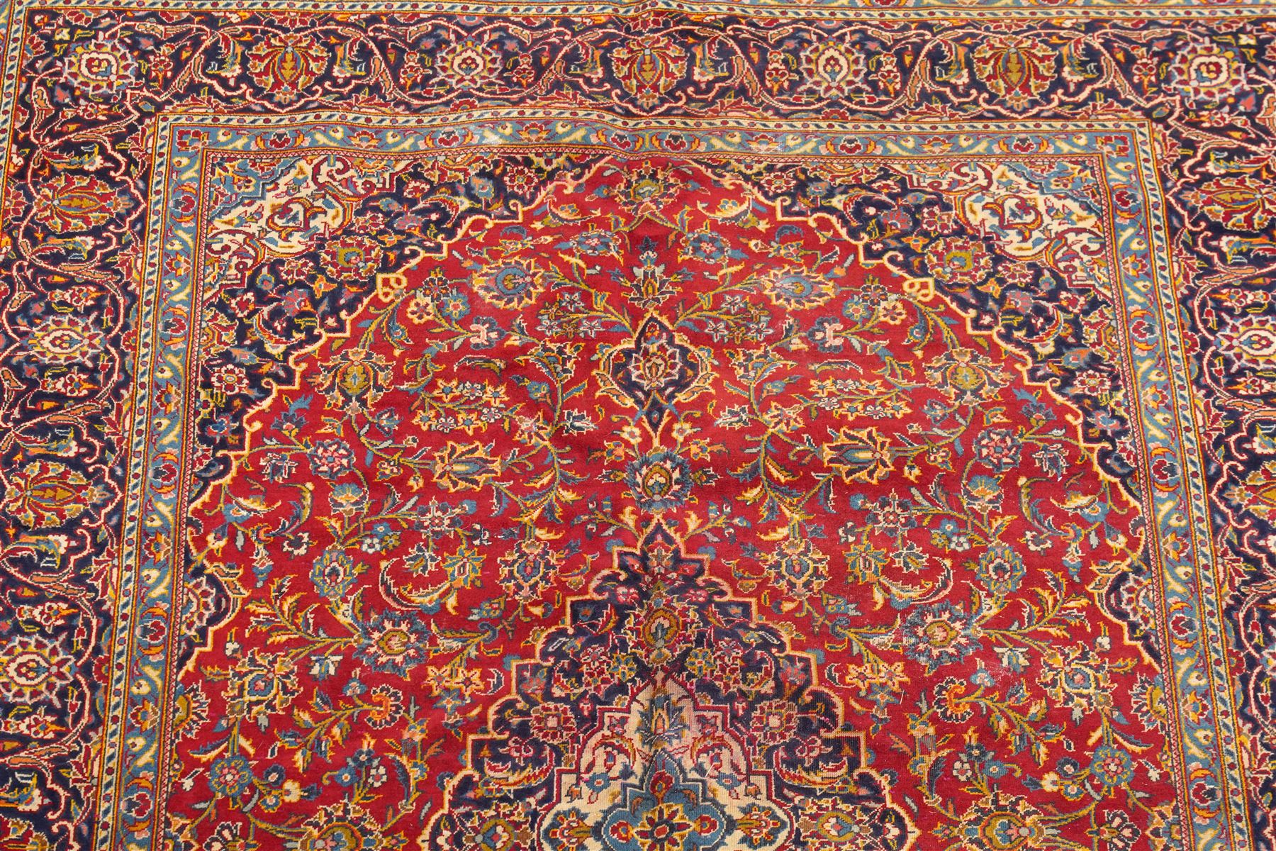 Persian Kashan hand-knotted carpet, madder red ground centred by shaped dark navy, ivory and pale blue floral medallion with pendants, surrounded by scrolling vines, palmettes and flowerheads, dark navy spandrels, enclosed by dark navy floral border with multiple floral guard stripes