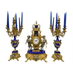 A 20th century continental gilt metal Lyre mantle clock with a pair of matching six light candelabra, spring driven movement housed in a gilt drum case on a brass and cobalt blue base with paw feet, two bronzed metal figures of mythological cherubs with animal legs and cloven hooves supporting a festooned garland swag, eight-day twin barrel striking movement with a floating lever balance escapement, striking the hours and half-hours on two bells, white enamel dial with roman numerals, minute track and pierced steel hands, dial inscribed “Imperial”. 