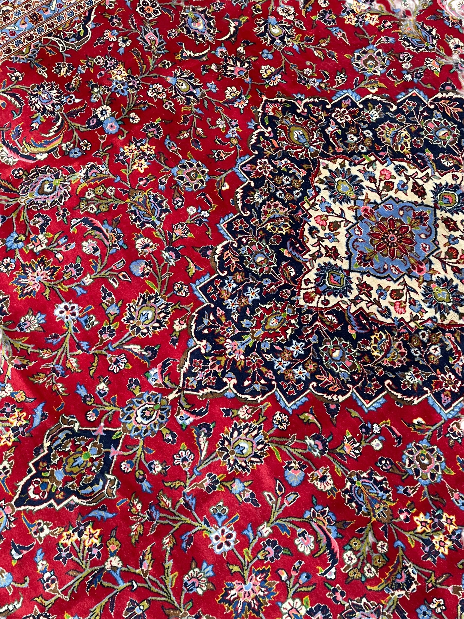 Persian Kashan crimson ground carpet, floral pattern indigo ground central medallion and matching spandrels, the field decorated densely with scrolling foliate patterns and floral motifs, the main border decorated with scrolling branches and palmettes, within floral pattern guard stripes