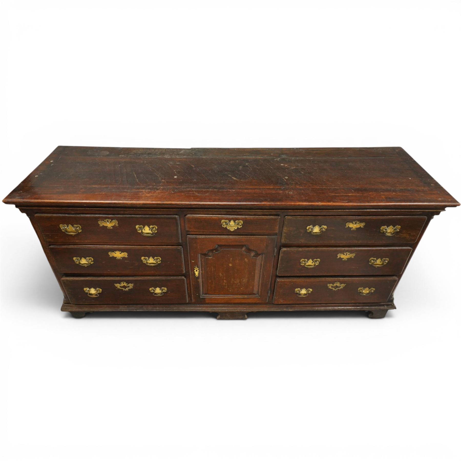 19th century Georgian oak long dresser base, rectangular plank top above a central frieze drawer over a fielded panel cupboard, flanked by three banks with quarter cantered pilasters, brass swan neck handles and escutcheons, raised on shaped bracket feet