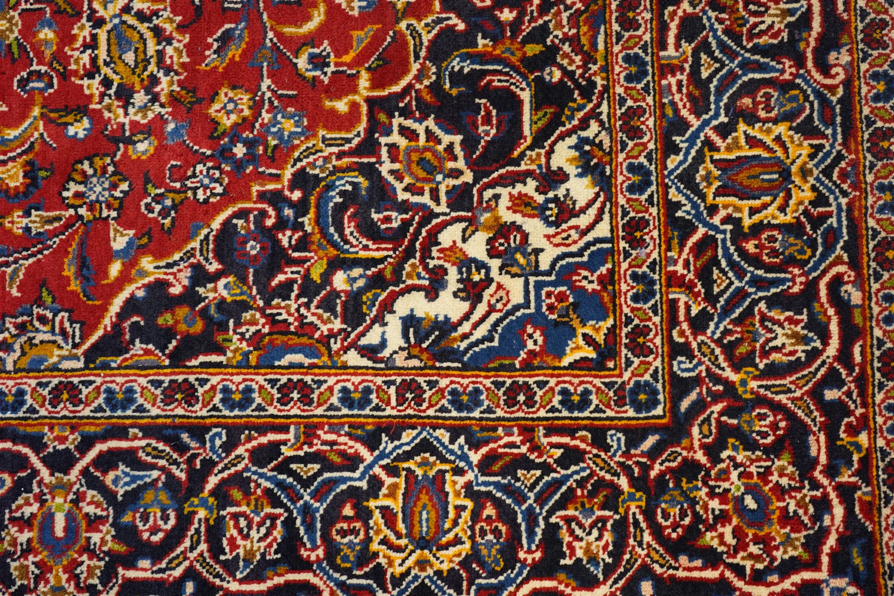 Central Persian Kashan hand-knotted rug, madder red field, with central lobed medallion with pendants, issuing an all-over design of scrolling vine, palmettes, and floral sprays, with conforming spandrels, enclosed by a broad indigo palmette and vine border framed by guard stripes in a palette of navy, blue and ivory tones, with cotton fringe ends 