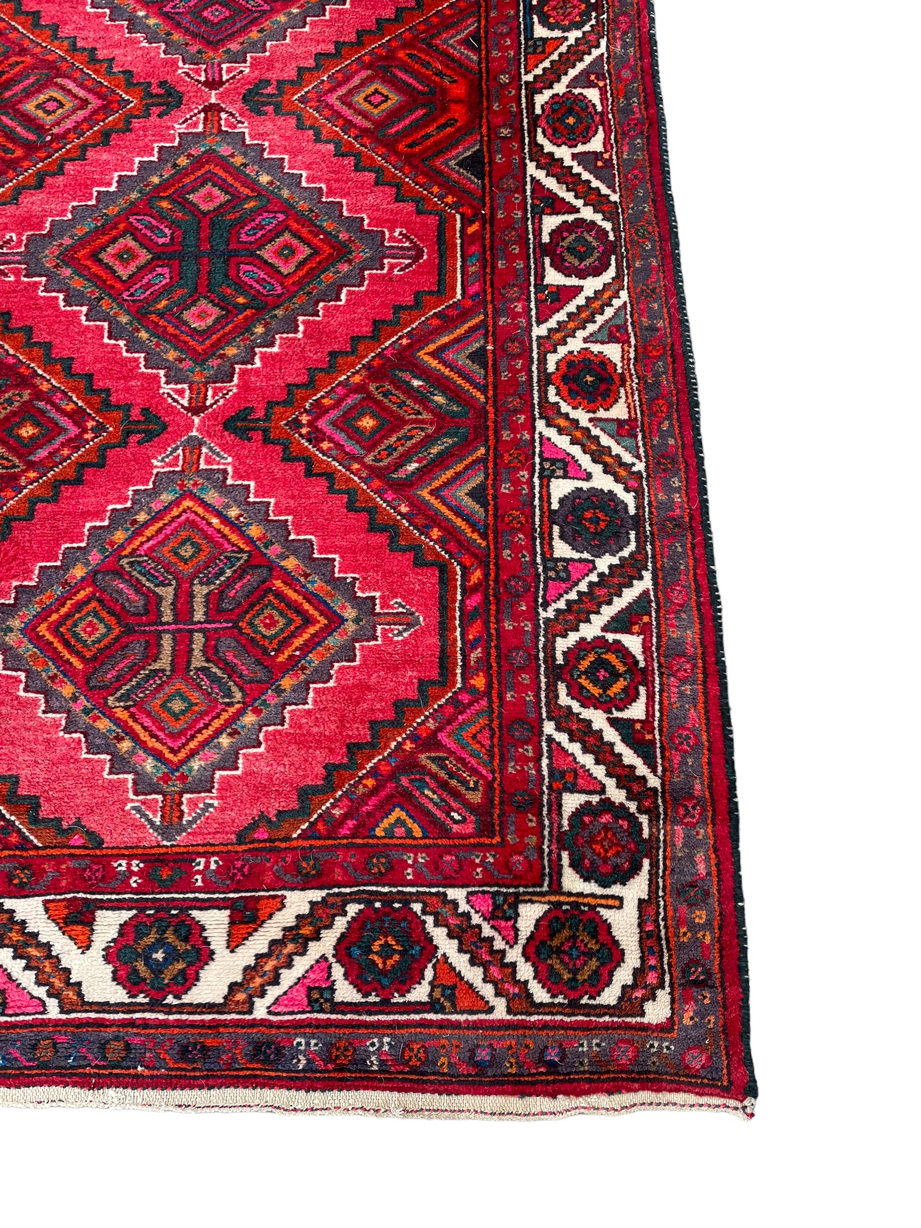 Persian Hamadan red ground rug, decorated with six stepped lozenge medallions, the guarded border with repeating stylised flower head decoration, with inscription to corner