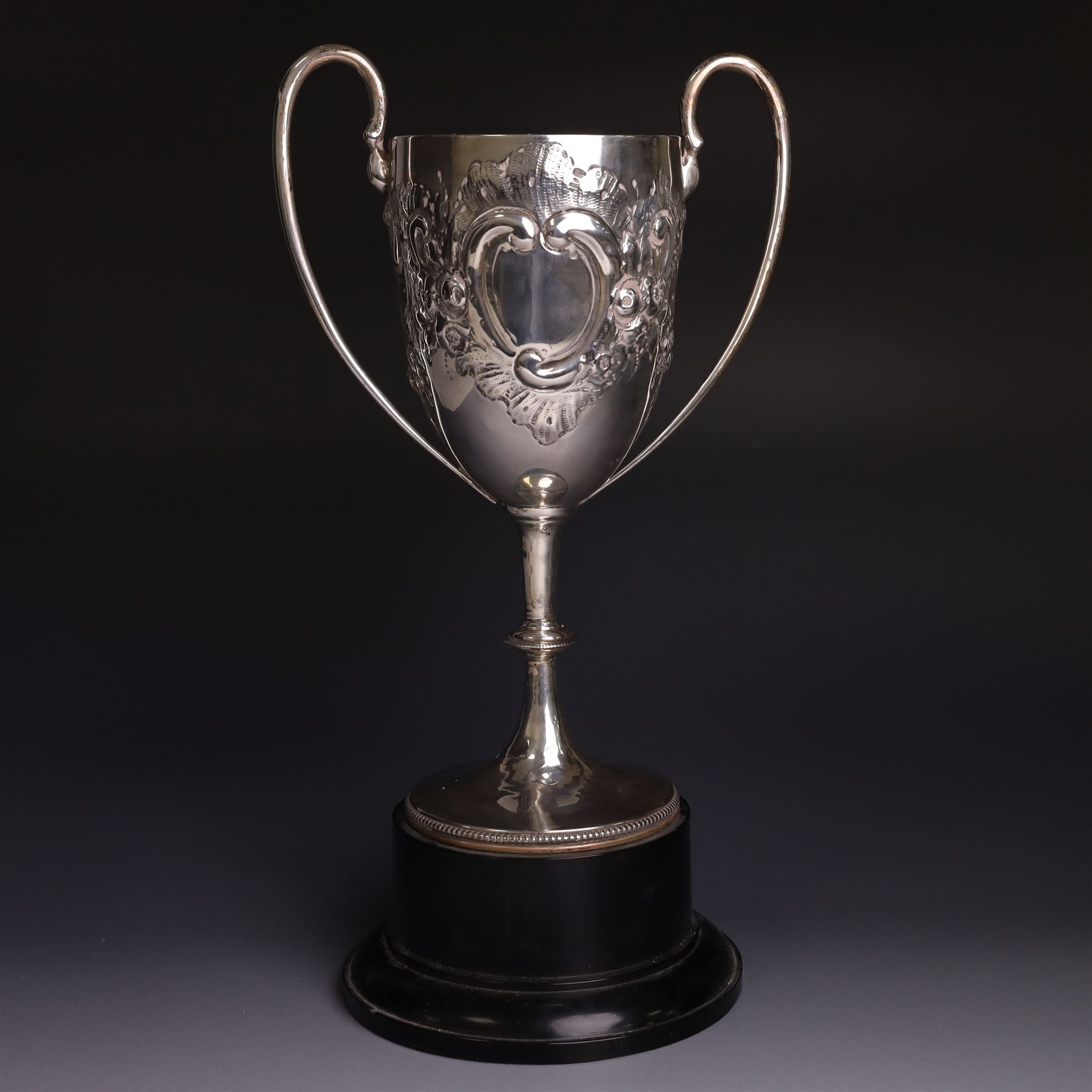Edwardian silver twin-handled trophy, with embossed floral and scrolling decoration, upon a knopped stem and circular foot, hallmarked Goldsmiths & Silversmiths Co Ltd, London 1903, upon a black plinth, including plinth and handles H32cm