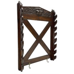 Victorian oak wall hanging whip or stick rack, sloped arch pediment carved with central grotesque mask with extended scrolling foliage, the upright supports cut with rests, x-framed back, two lower hooks