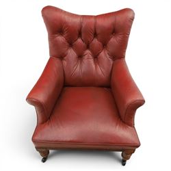 Early 20th century style oxblood red leather Chesterfield armchair, deep buttoned shaped back over padded scroll arms and loose seat cushion, raised on turned mahogany front supports with brass caps and castors