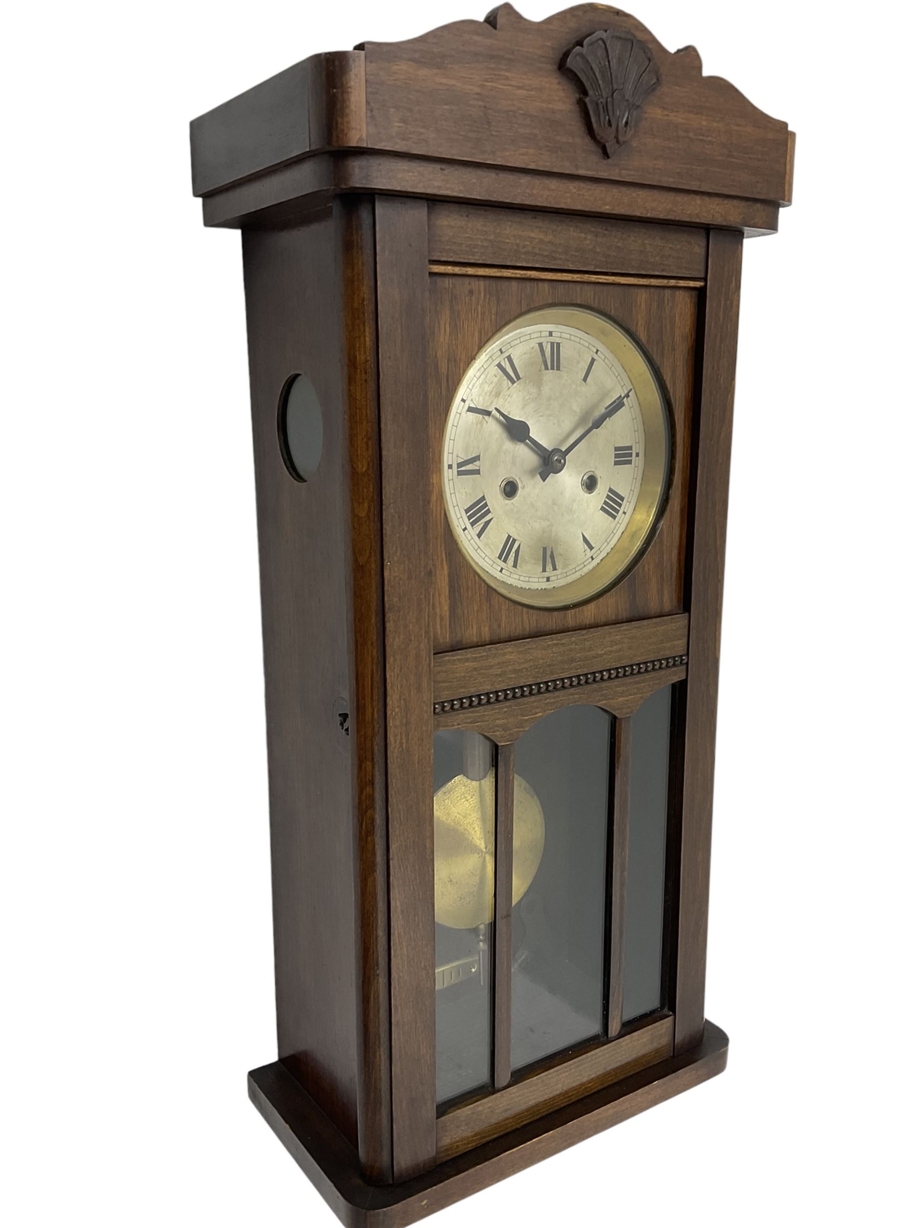1930s wall clock in an oak case with a crested pediment and glazed door with visible pendulum, silvered dial with Roman numerals and steel spade hands, eight day spring driven movement striking the hours and half hours on a gong. With pendulum and key.