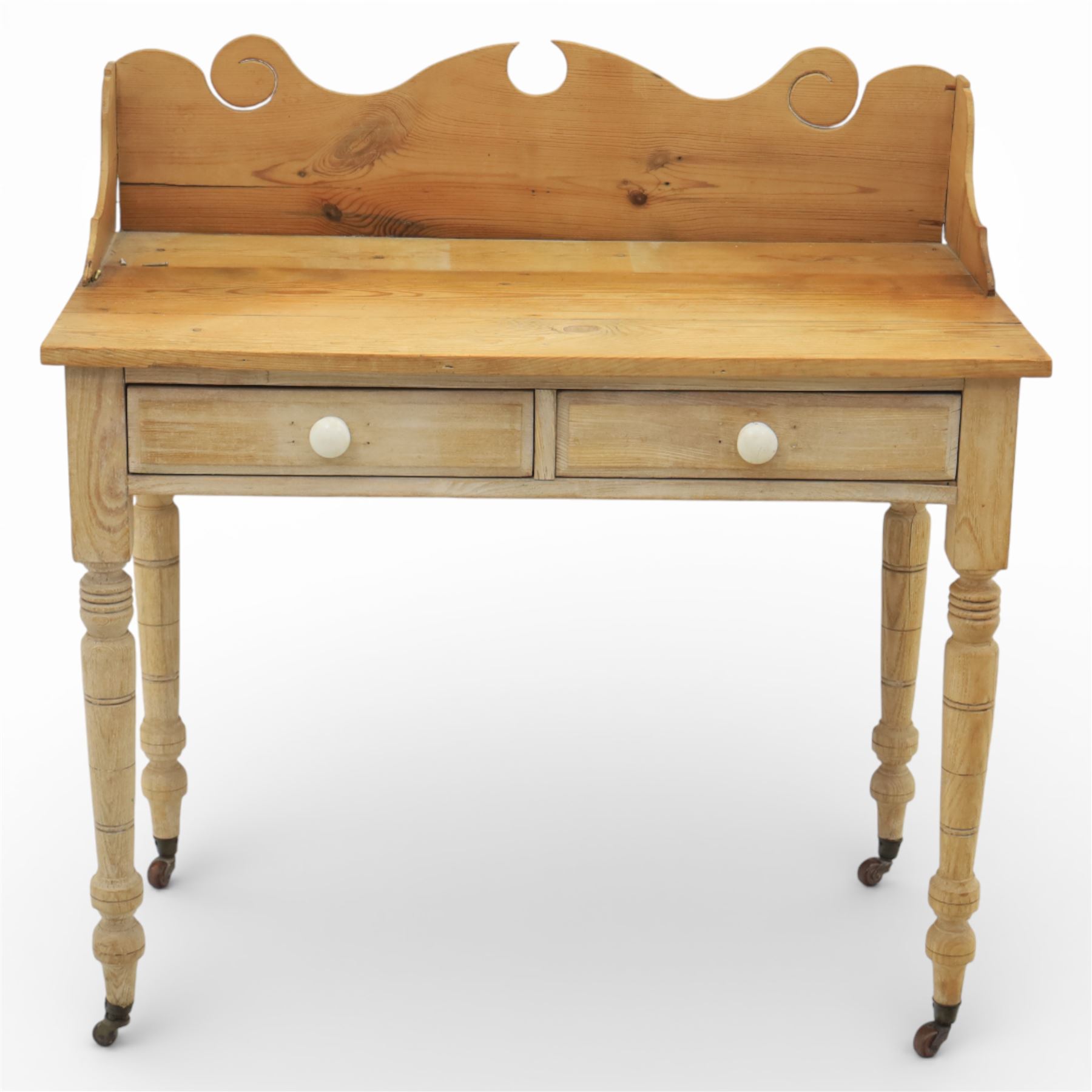 19th century stripped pine side table, the rectangular plank top with low shaped back rail incorporating scroll cut ends, fitted with two frieze drawers with ceramic knob handles, raised on turned supports terminating in castors