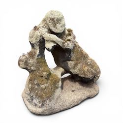 Carved composite stone garden figure, depicting a seated child with folded arms, weathered finish with moss and lichen growth, raised on an integral naturalistic base
