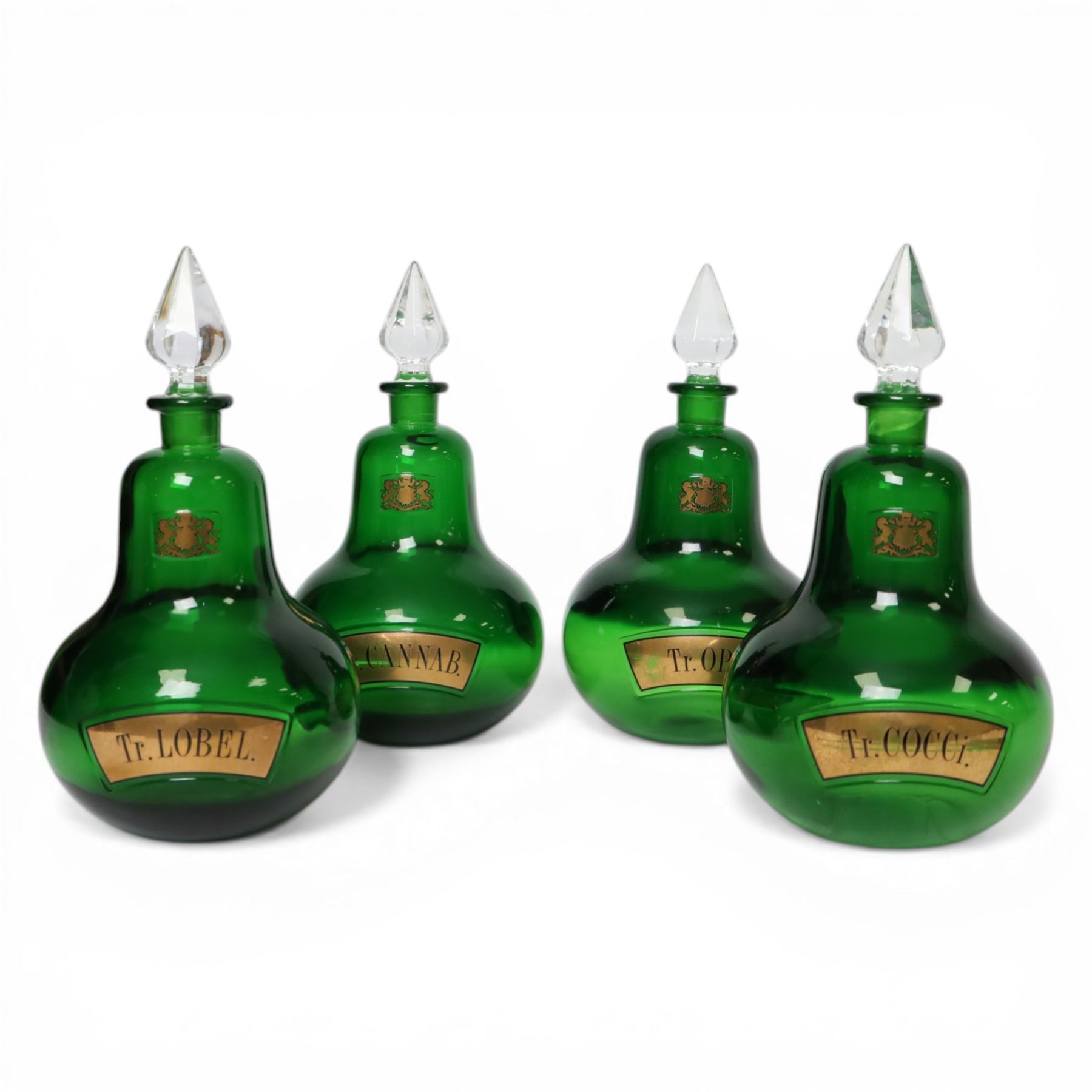 Set of four 20th century Royal Pharmaceutical Society green glass bottles, of bellied form with clear cut glass spire stopper, each body bearing gilt label in Latin: Tr.OPii., Tr.COCCi., Tr.CANNAB., and Tr.LOBEL., and gilt Royal Pharmaceutical Society crest to neck, H25cm

Provenance: From the collection of the family of Peter Green, a practising pharmacist whose career spanned the mid to late 20th century. During his professional life he managed pharmacies in Yeovil, Somerset, and later Selles Chemist in Filey, North Yorkshire, before purchasing and running his own business, Peter Green Pharmacy, located in the Market Place, Malton. The bottles were displayed as part of the traditional shop fittings in both the Filey and Malton pharmacies and were retained as part of his collection.