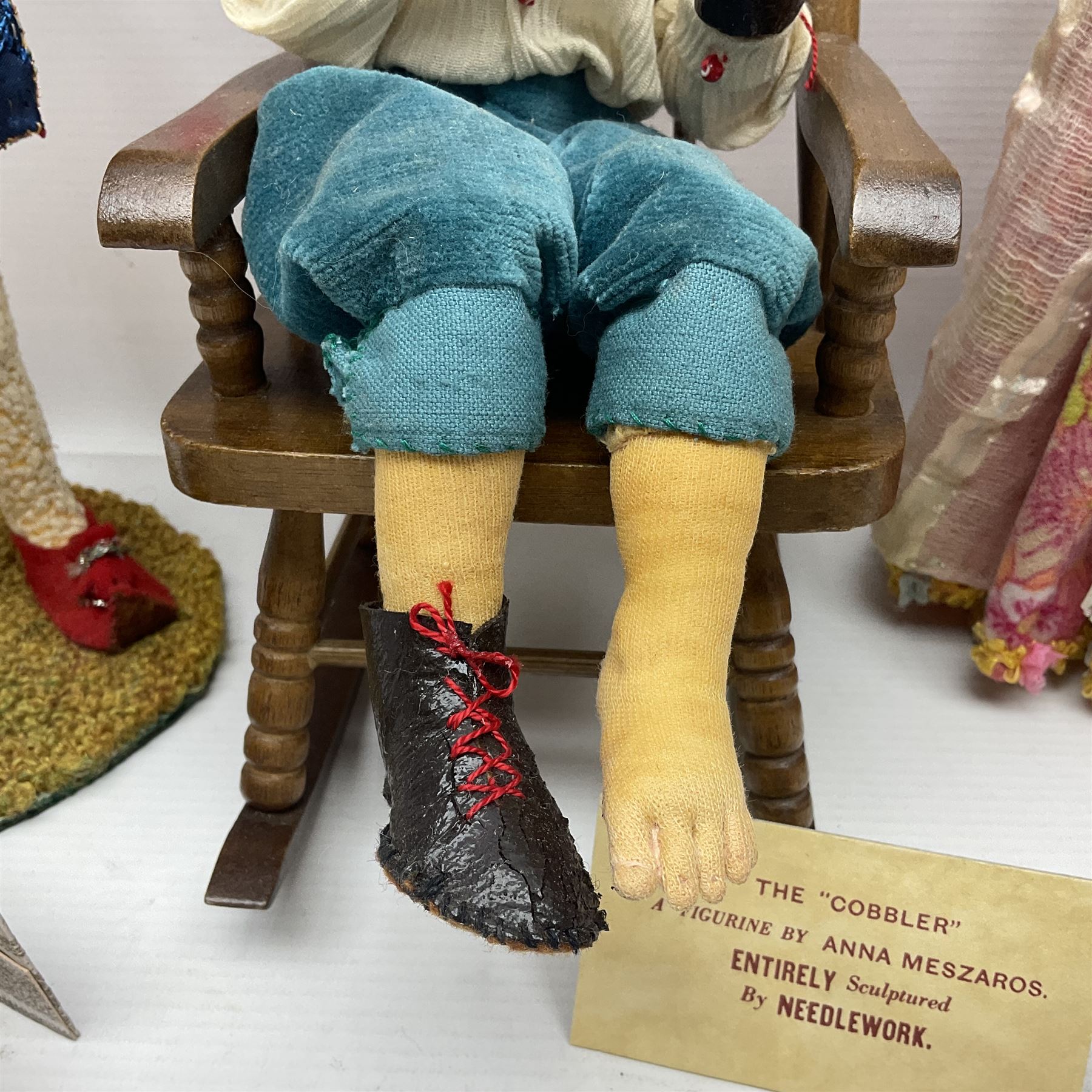 Anna Meszaros Hungary - three hand made needlework figurines - 'The Cobbler' as a child seated in a wooden rocking chair working on a removed boot H28cm; young girl in a long floral dress standing playing a harp H35cm; and young girl wearing a blue tabard standing playing a lute (3) Auctioneer's Note: Anna Meszaros came to England from her native Hungary in 1959 to marry an English businessman she met while demonstrating her art at the 1958 Brussels Exhibition. Shortly before she left for England she was awarded the title of Folk Artist Master by the Hungarian Government. Anna was a gifted painter of mainly portraits and sculptress before starting to make her figurines which are completely hand made and unique, each with a character and expression of its own. The hands, feet and face are sculptured by layering the material and pulling the features into place with needle and thread. She died in Hull in 1998.  