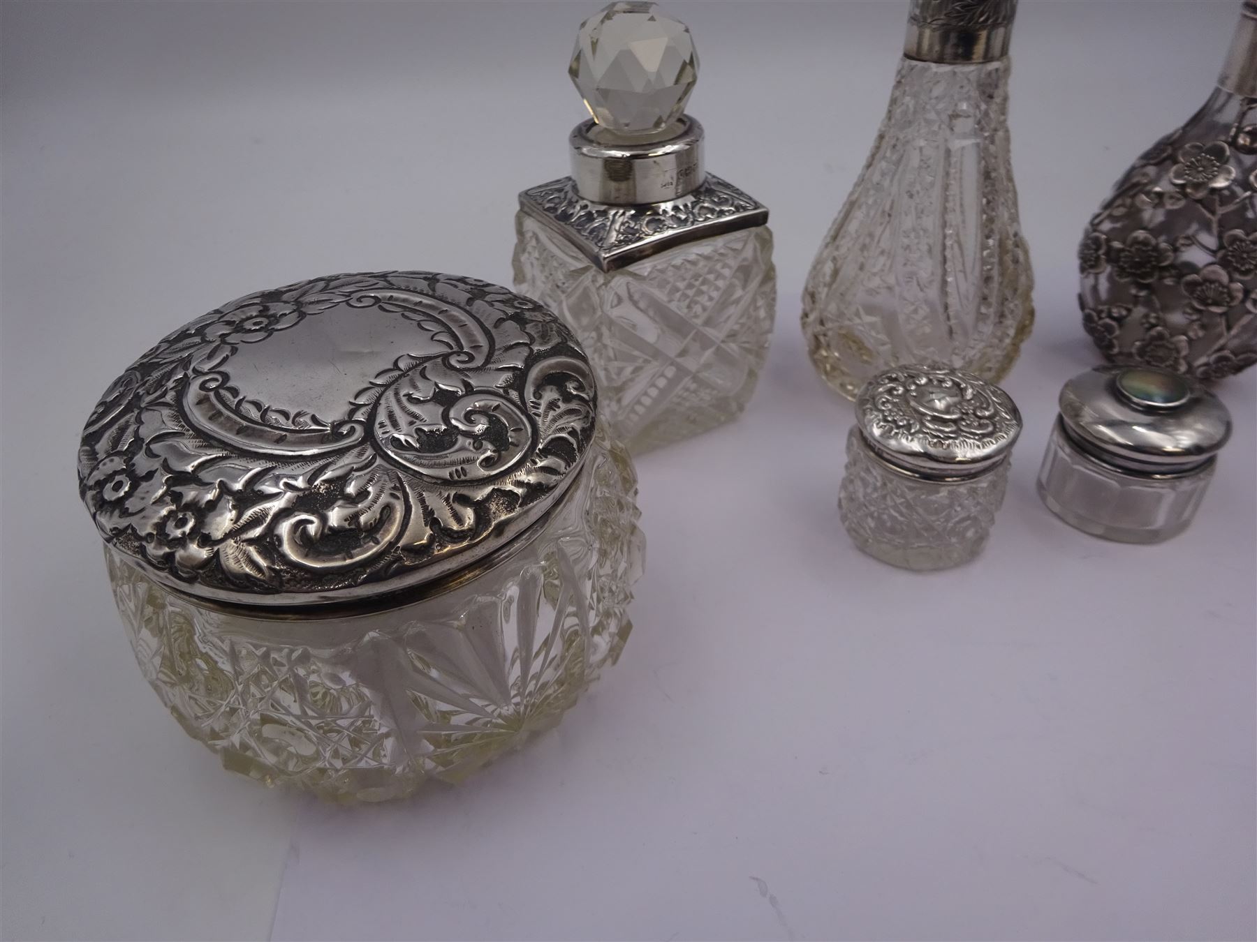 Two silver mounted dressing table jars, one in the form of a heart, both with embossed silver covers and cut glass bodies, hallmarked, together with two smaller examples, one set with mother of pearl, three silver mounted glass scent bottles, including an example with prunus blossom decoration, stamped Sterling, tallest bottle H17cm