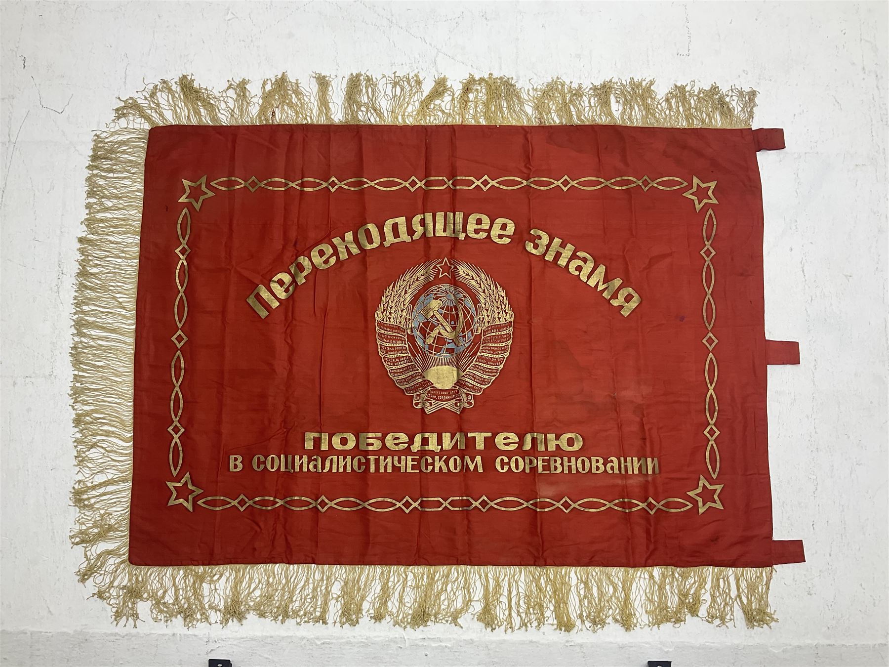 1970s Soviet banner printed in gold on a red ground, roughly translates as 'Transferable Banner - To the winner in the Socialist Competition' and 'Work Study Live by Communism' verso; wreath of wheat ears to either side; tassels on three sides 110 x 155cm