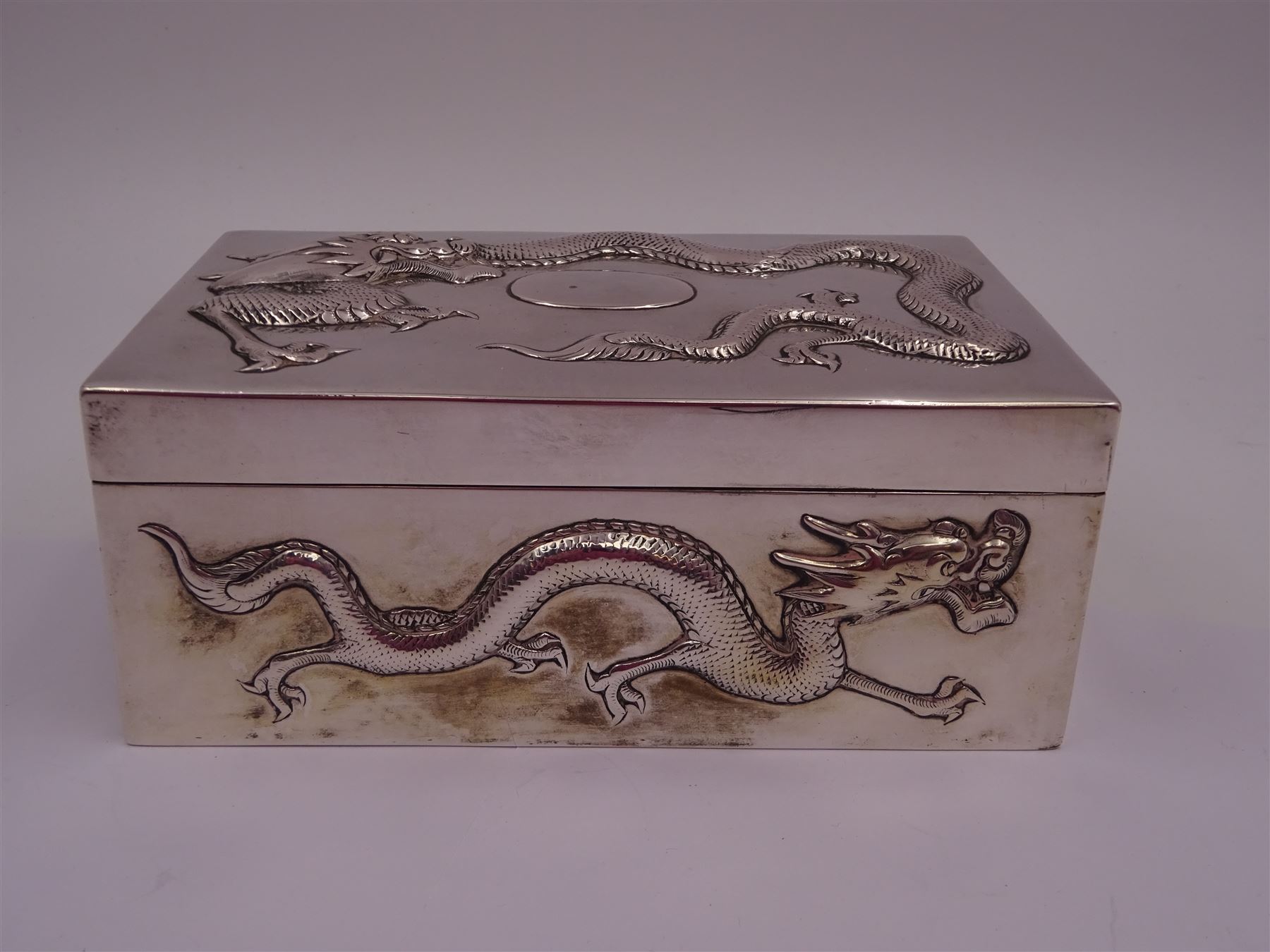 Late 19th century/early 20th century Chinese export silver mounted cigarette box, of rectangular form, decorated in relief with dragons, the hinged cover with similar dragon decoration and central blank oval cartouche, opening to reveal a softwood lined compartmentalised interior, stamped beneath with character mark and KMS for maker Kwong Man Shing, H6.3cm, W15.5cm, D9.5cm