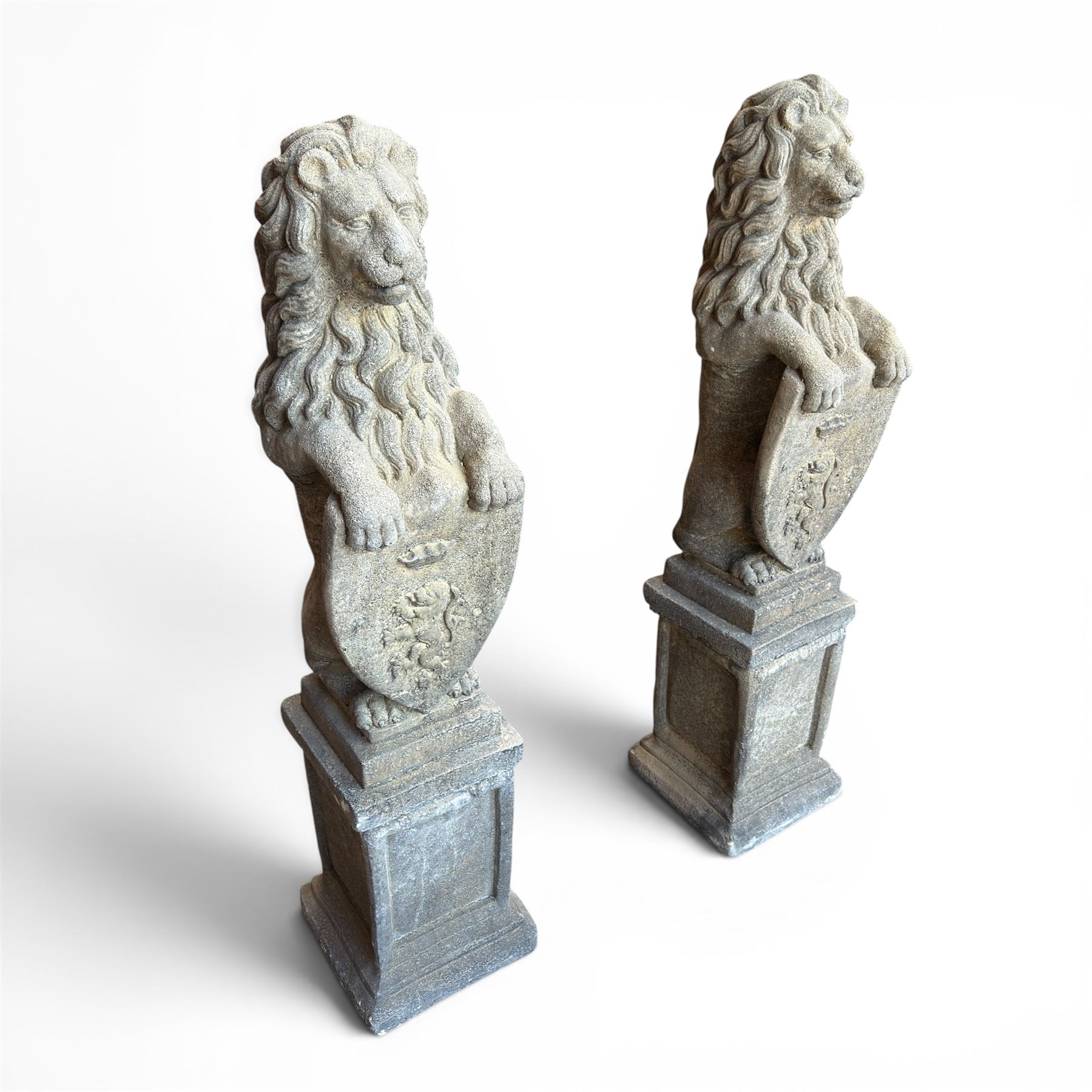 Pair of cast stone Heraldic lions, modelling in upright standing poses with flowing mane, each clasping an Armorial shield moulded with a crowned lion in their forepaws, raised on rectangular pedestals