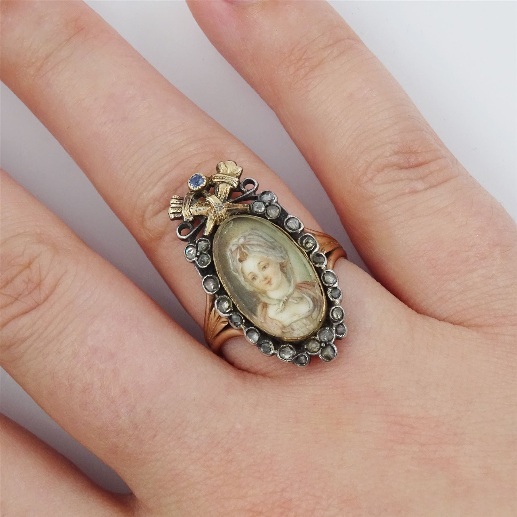 18th / 19th century silver and gold diamond and sapphire portrait ring, set with an oval miniature of a woman wearing a bonnet on ivory, under a glazed panel, surmounted with a 'trophy of love' symbol, modelled as a flaming torch and bow and quiver, set with a single round cut sapphire and rose cut diamond surround, to trifurcated shoulders, on French 18ct gold shank, Eagle hallmark. This lot has been registered for sale under Section 10 of the APHA Ivory Act