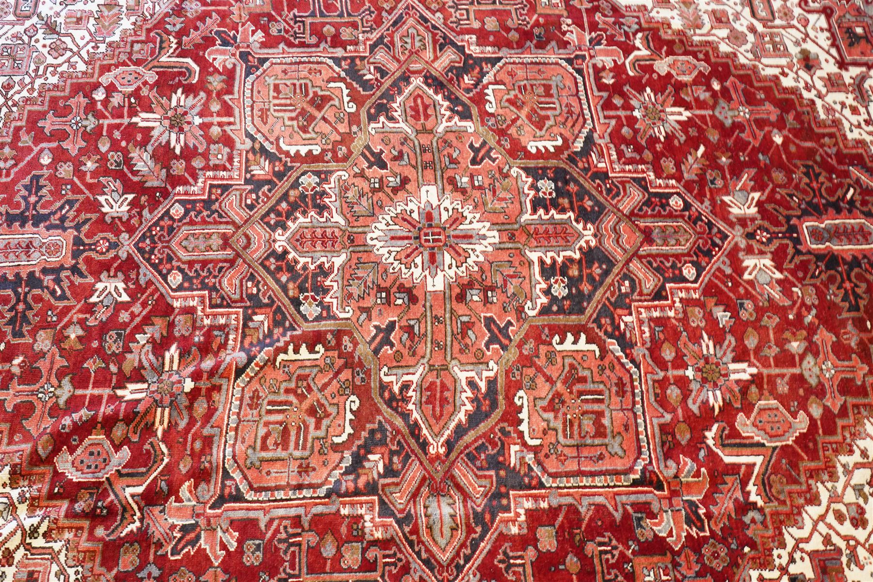 North West Persian Heriz hand-knotted carpet, brick red field with a large central angular star medallion with pendants surrounded by stylized geometric palmettes, serrated leaves and tribal motifs,, ivory corner spandrels, field enclosed by a dark indigo main border of stylize rosettes and angular vine motifs between multiple guard stripes, with cotton fringe ends 