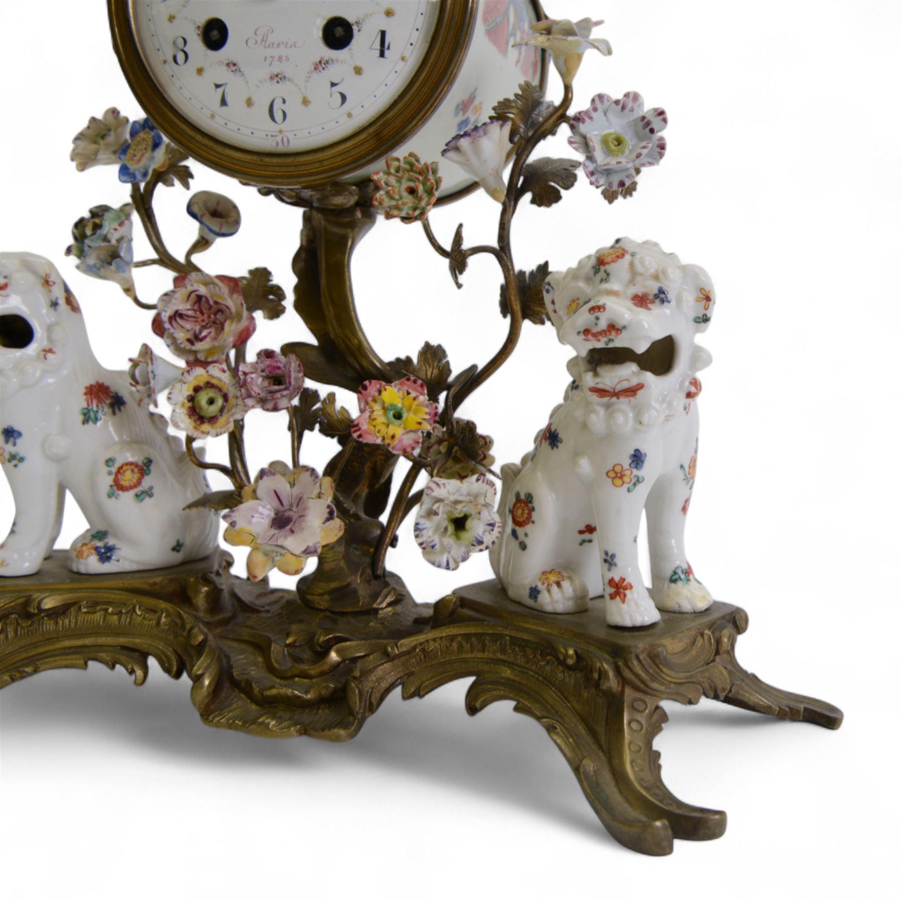 French- early 20th century 8-day Chinoiserie brass and porcelain mantel clock in the 18th century style, brass base with splayed feet profusely decorated with clusters of painted porcelain flowers and two decoratively painted Chinese foo dogs, movement enclosed in a circular porcelain movement case illustrated with birds of paradise and oriental motifs, with a white enamel dial Arabic numerals, minute markers, pierced gilt hands and a floral swag, rack striking Parisian movement striking the hours and half-hours on a bell. With a visible sunburst pendulum and key.
