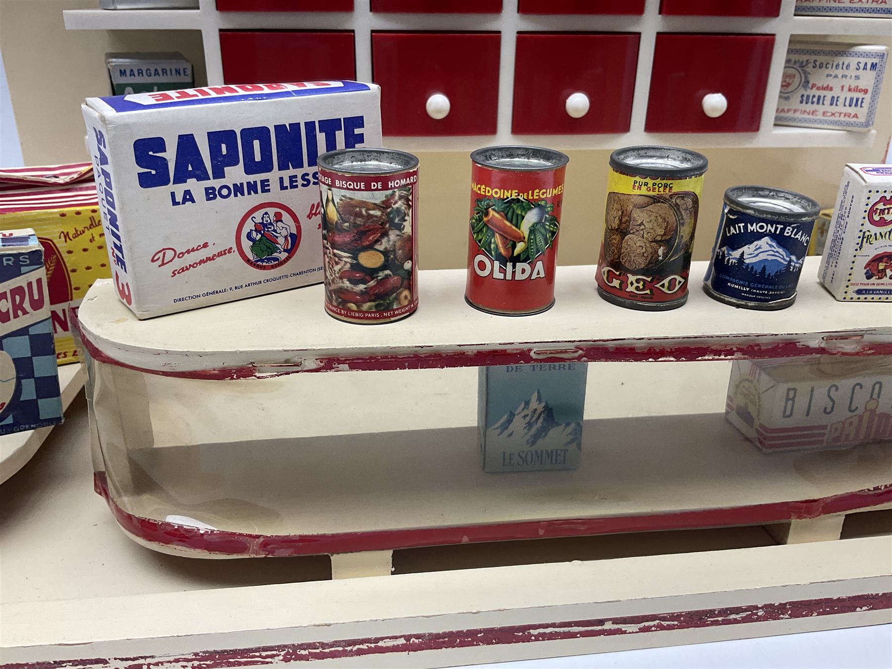 Late 1950s continental red and white painted wooden diorama of a grocery shop interior with fitted shelves and drawers and freestanding 'glazed' counter, fully stocked with miniature tin cans and packets of food L42cm H23cm D22cm
