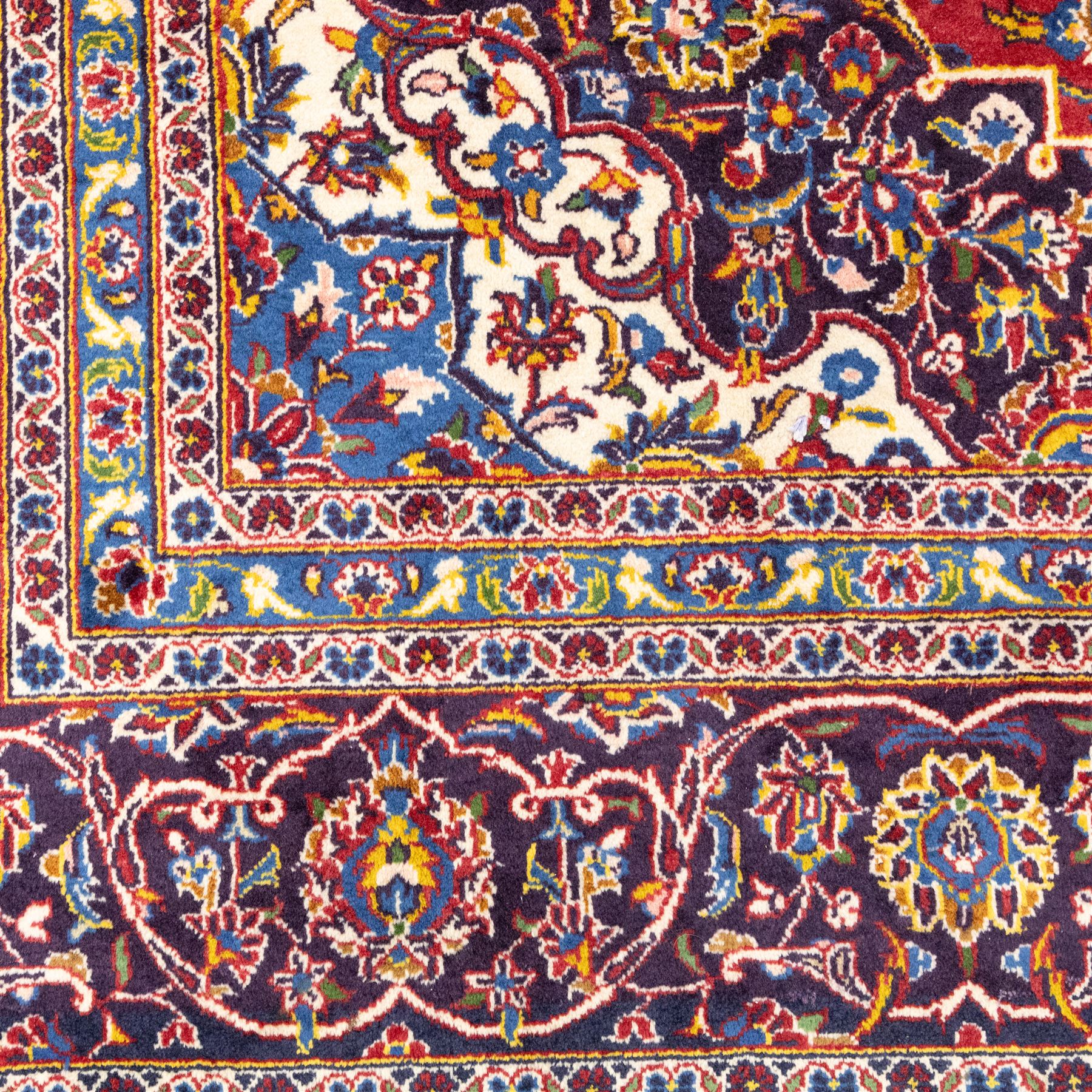 Central Persian Kashan hand-knotted carpet, crimson central field with lobbed medallion in navy blue and ivory ground, surrounded by a dense all-over floral design of scrolling vines, palmettes, and stylized blossoms, enclosed within a deep navy main border, flanked by multiple guard stripes with fringe ends 


