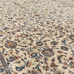 Large Central Persian Kashan hand-knotted carpet, ivory field with an all-over scrolling design of vines, palmettes, and floral sprays, enclosed by a broad camel-ground palmette and vine border, framed by multiple guard stripes in a palette of ivory, beige, blue and polychromed tones, with cotton fringe ends 