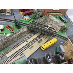 Table top 'N' gauge continental scenic layout as a split level town with various loops of track, station with numerous platforms, engine shed, assorted buildings, roads with motor vehicles and figures, tunnels, trees, river with bridge, tram depot etc 122 x 52cm
